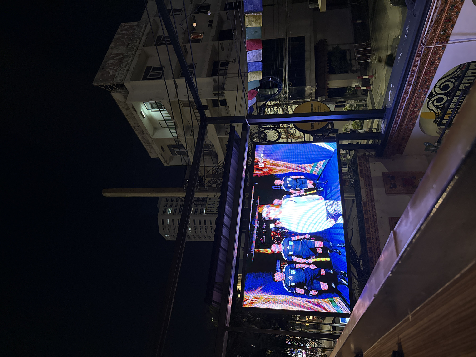 The image depicts an outdoor nighttime scene in Bangkok, Thailand, viewed from an elevated position, possibly a balcony or rooftop. In the foreground, a portion of a wooden railing is visible. Dominating the mid-ground is a large, bright LED screen displaying a live broadcast of a football match. The screen shows several men in referee uniforms, distinguished by their black shirts and shorts with white accents, walking onto a field. The backdrop of the screen is a vibrant blue with abstract patterns.
Behind the screen, several buildings are visible. To the right, a multi-story building with a weathered facade has the faded sign "PAN PAN" at the top. Below this, a lighter-colored building with multiple windows and balconies is illuminated. Various wires and cables are strung across the scene. Prayer flags in multiple colors are hung horizontally across a black metal frame, adding a cultural element.
Further in the background, a tall, slender, light-colored structure, possibly a smokestack or utility tower, rises against the dark night sky. Another tall, modern building with many windows is visible behind it. The environment appears to be an urban setting with street-level activity implied by the visible street and some light poles. The overall impression is a lively urban night atmosphere, likely a viewing area for a sporting event.
