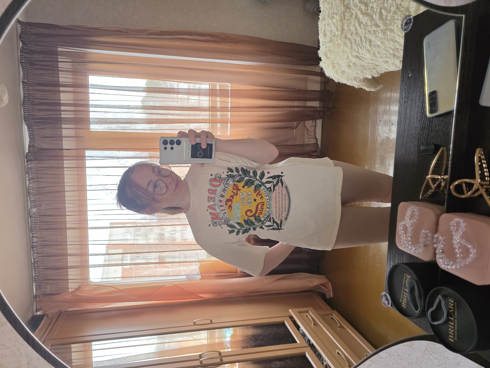 A young woman with glasses is taking a selfie in a mirror. She is wearing a light-colored t-shirt with a graphic design. The mirror reflects a room with a window, curtains, and a building visible outside. Next to the mirror is a shelf or table with accessories, including boxes and what appears to be eyewear. A phone is lying on the table. The image was taken in an apartment in Chaikovsky, Russia, at a location with windows overlooking a residential building.