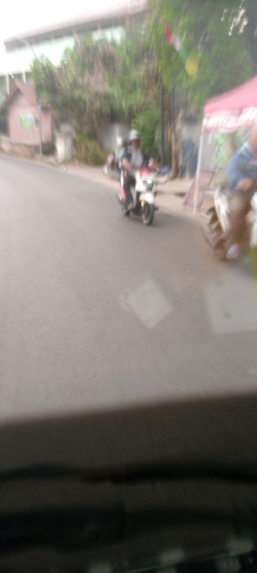 The image depicts a blurry street scene with several motorbikes and people. In the foreground, the edge of a dark interior, likely a vehicle, is visible. The middle ground shows a street with at least two motorbikes in motion. One white and red motorbike is prominently featured, ridden by a person wearing a helmet and a grey shirt. To the right, another motorbike is partially visible, ridden by a person in a blue shirt. In the background, there are buildings, trees, and a pink tent. The overall impression is one of movement and a casual outdoor setting, possibly in a residential or commercial area. The presence of motorbikes suggests an urban environment. Based on the context of the provided location information, this scene is likely occurring in Bandung City, Indonesia.