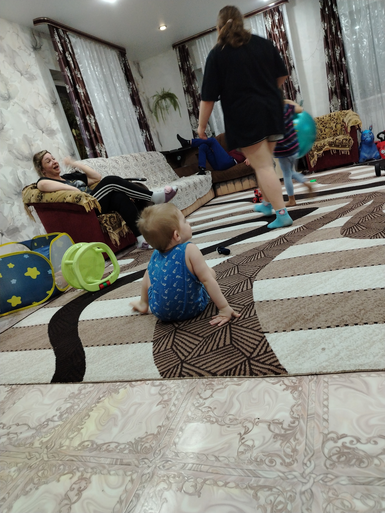 The image depicts an interior scene in what appears to be a home in Большая Уса, Russia. A blonde-haired woman reclines on a couch, wearing a black top and black pants with white stripes, engaged in conversation or laughter. In the foreground, a baby with light hair sits on a patterned rug, wearing a blue sleeveless shirt with a printed design. Beside the baby is a small, blue and yellow toy tent and a green toy.
A woman in a black t-shirt and shorts walks across the room, followed by a child in blue socks holding a blue balloon. A tan couch and a maroon upholstered chair are visible in the background, along with patterned curtains hanging in front of a window. The floor is tiled with patterned flooring extending from underneath the rug. There are toys and other objects around the room.
