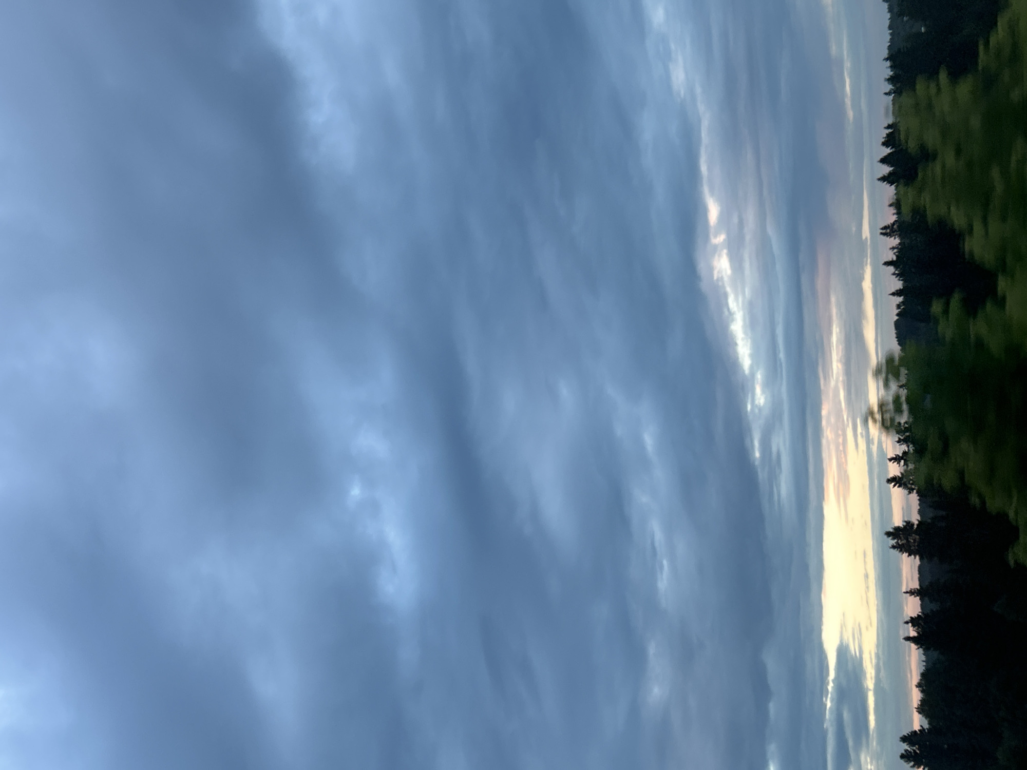 The image shows a sky filled with dark, heavy clouds. Below the clouds, a line of dark green coniferous trees stretches across the horizon. A band of soft, warm light is visible in the sky just above the trees, suggesting either sunrise or sunset. The overall atmosphere is moody and overcast. The location is identified as Забегалово, Russia. There are no people or visible objects in the scene other than the natural environment.
