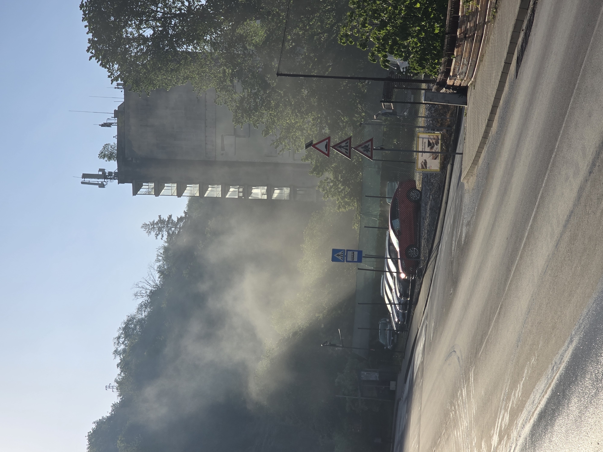 The image captures an urban or semi-urban street scene enveloped in a significant amount of mist or smoke, dramatically illuminated by sunlight. A wide, asphalt road, appearing wet or recently damp, occupies the right side of the frame, reflecting the bright light. A concrete sidewalk borders the road, separated from a dense, tree-covered hillside on the left by a low fence.
A multi-story, grey concrete building stands prominently on the far left, its lower floors partially obscured by trees and the pervasive mist. The building features large, reflective windows, and its rooftop is equipped with multiple antennas and communication structures.
Along the right side of the road, at least two cars are parked, with a red car most prominent in the mid-ground. Several traffic signs are visible: three red-bordered triangular warning signs, and a blue square sign indicating a pedestrian crossing, possibly with a bus stop symbol or the word "СПИРКА" (Stop/Bus Stop) below it. A large billboard, partially hidden by foliage and mist, displays an advertisement.
The low angle of the sunlight, shining from the right, suggests either early morning or late afternoon. This light dramatically illuminates the dense mist, giving it a bright, ethereal quality as it rises from or covers the hillside and the lower parts of the buildings and trees. No human activity is discernible.
Visible text on the billboard, in Cyrillic script, includes "Магазин за мъжко облекло" (Store for men's clothing).