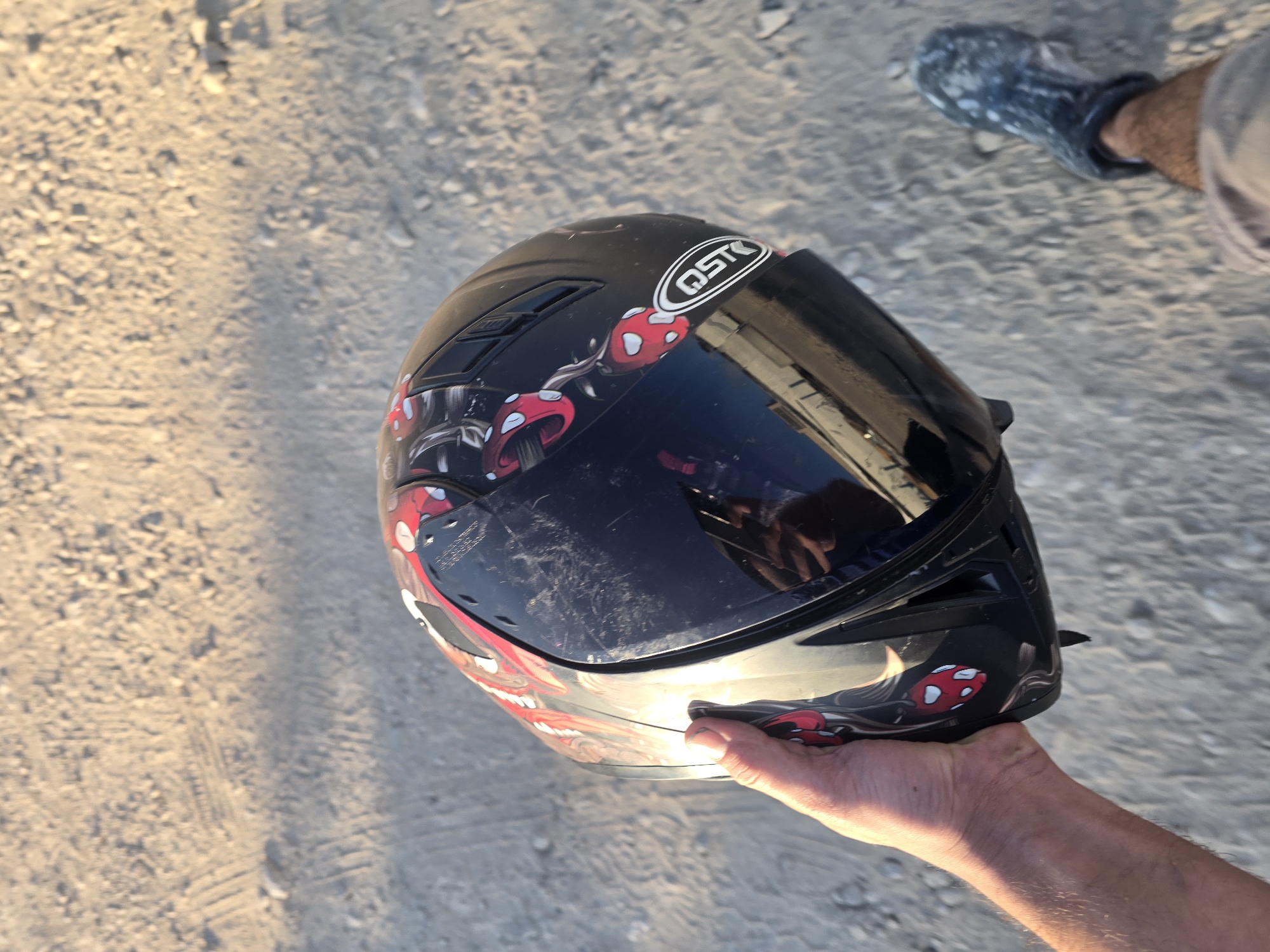 A person's hand holds a black motorcycle helmet with a red and white mushroom design and the "QST" logo. The helmet has a clear visor, reflecting a portion of the scene. The person is wearing light-colored shorts and a patterned boot on their right foot. The ground appears to be a dusty or sandy surface with some tire tracks. The lighting suggests it is daytime. The location is Yambol, Bulgaria.