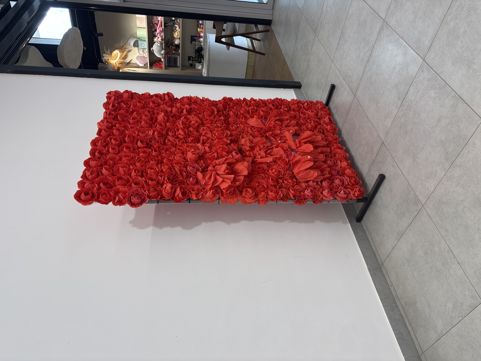 An eye-level, medium shot captures a large, rectangular floral installation, composed entirely of vibrant red paper roses, positioned against a plain white wall. The installation is supported by a black metal stand with horizontal supports at the base. To the right of the floral display, a doorway opens into a brightly lit store interior. The store features shelves stocked with various items, including plush toys, decorative boxes, and mugs. A white counter and a wooden chair are also visible within the store. The floor is tiled with large, light gray square tiles, and a grey baseboard runs along the wall. The scene is devoid of people. The overall lighting suggests it is daytime.