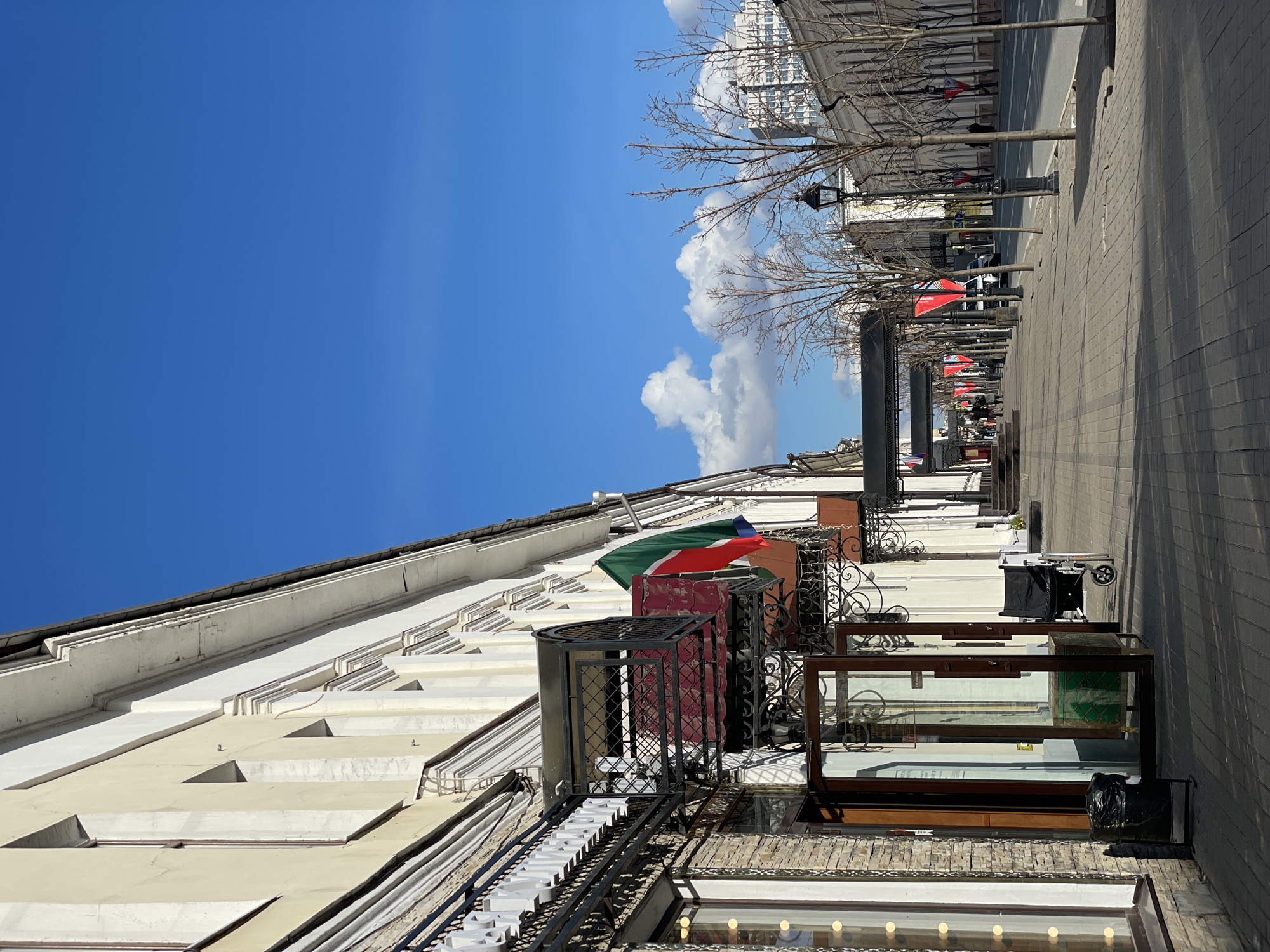 This image presents a sunny, ordinary street scene in a city, consistent with the provided context of Kazan, Russia. A row of multi-story buildings with light-colored facades and classic architectural details lines the left side of the frame. Many of these buildings feature ornate black wrought iron balconies or railings, and their ground floors appear to house shops with large glass display cases.
On the right, a wide, paved sidewalk stretches into the distance, paralleled by a street that is partially visible. The sidewalk is lined with several bare deciduous trees and streetlights, suggesting either late winter or early spring. The sky above is a vibrant blue with scattered white cumulus clouds, indicating clear and bright weather, with strong sunlight casting distinct shadows.
Two flags are visible: one prominently displaying horizontal bands of green, red, and blue (consistent with the Flag of Dagestan) hangs from a balcony on one of the buildings. Further down the street, another predominantly red banner or flag can be seen. A few indistinct figures of people are visible walking on the distant sidewalk, engaging in typical daily urban activity.
Visible text includes white lettering on a shop sign, partially readable as "МИНИСТ" (MINIST). A faint white "STOP" sign shape is also discernible on a distant red banner. The scene conveys a calm and active urban environment.