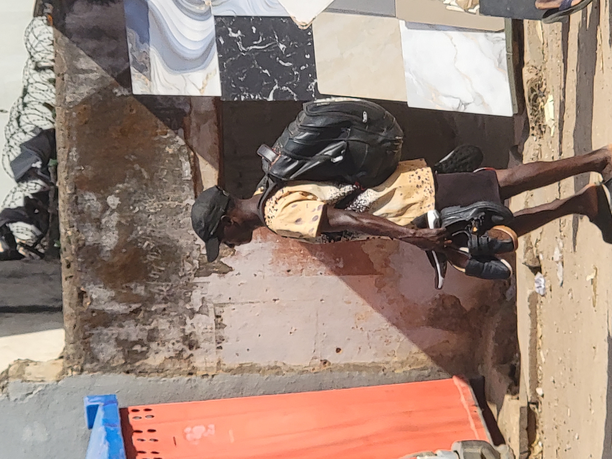 A man is lying on the ground in Maiburiji, Nigeria. He is wearing a black cap, a yellow patterned shirt, and black shorts. He has a black backpack on his back and is wearing black shoes. He is lying on a sidewalk next to a building with tiled walls. There is a red and blue object in the foreground. The scene is well-lit with strong shadows.