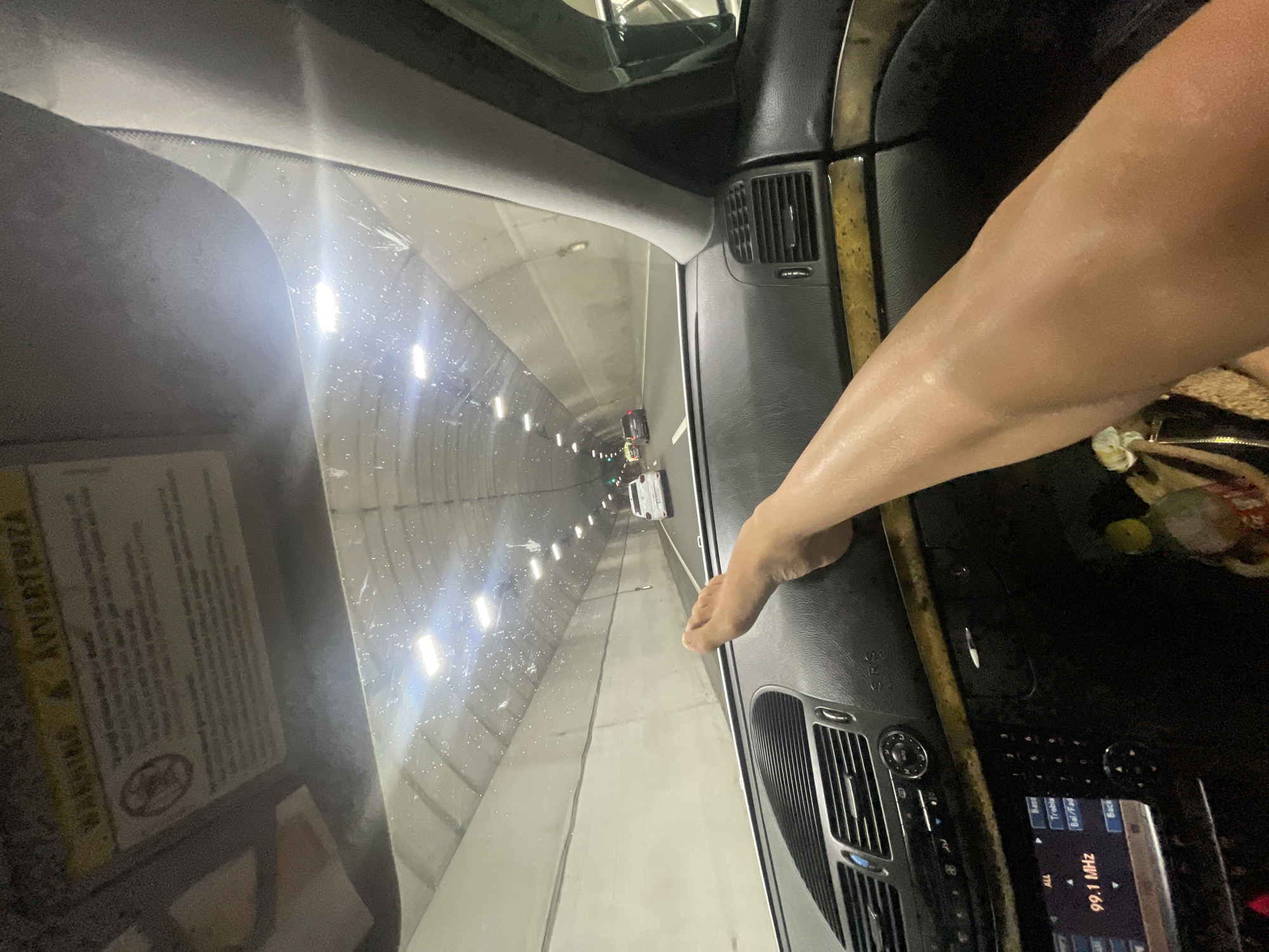 An interior view from a vehicle in Golemo Buchino, Bulgaria, shows a bare, tan-skinned leg and foot resting horizontally on the black dashboard. The foot is positioned above the central air vents, with the leg extending towards the lower right of the frame. The car's dashboard features silver-colored air vent grilles and control dials. Below the dashboard, a media system display shows "99.1 MHz" and menu options including "Bass," "Treble," "Bal/Fad," and "Back." A sun visor is visible in the upper left, displaying "WARNING" and "AVVERTENZA" text, along with a circular symbol that is partially obscured but appears to be a warning graphic.
Through the windshield, a brightly lit tunnel is visible. The tunnel walls are a light beige or grey, constructed from horizontal concrete segments. Numerous rectangular light fixtures are mounted overhead, illuminating the tunnel. The windshield surface displays multiple small specks, possibly water droplets or dust. In the distance, other vehicles are present within the tunnel, including a white car in the right lane. A green light, likely a traffic signal, is discernible further ahead in the tunnel. The view also encompasses the car's right A-pillar.