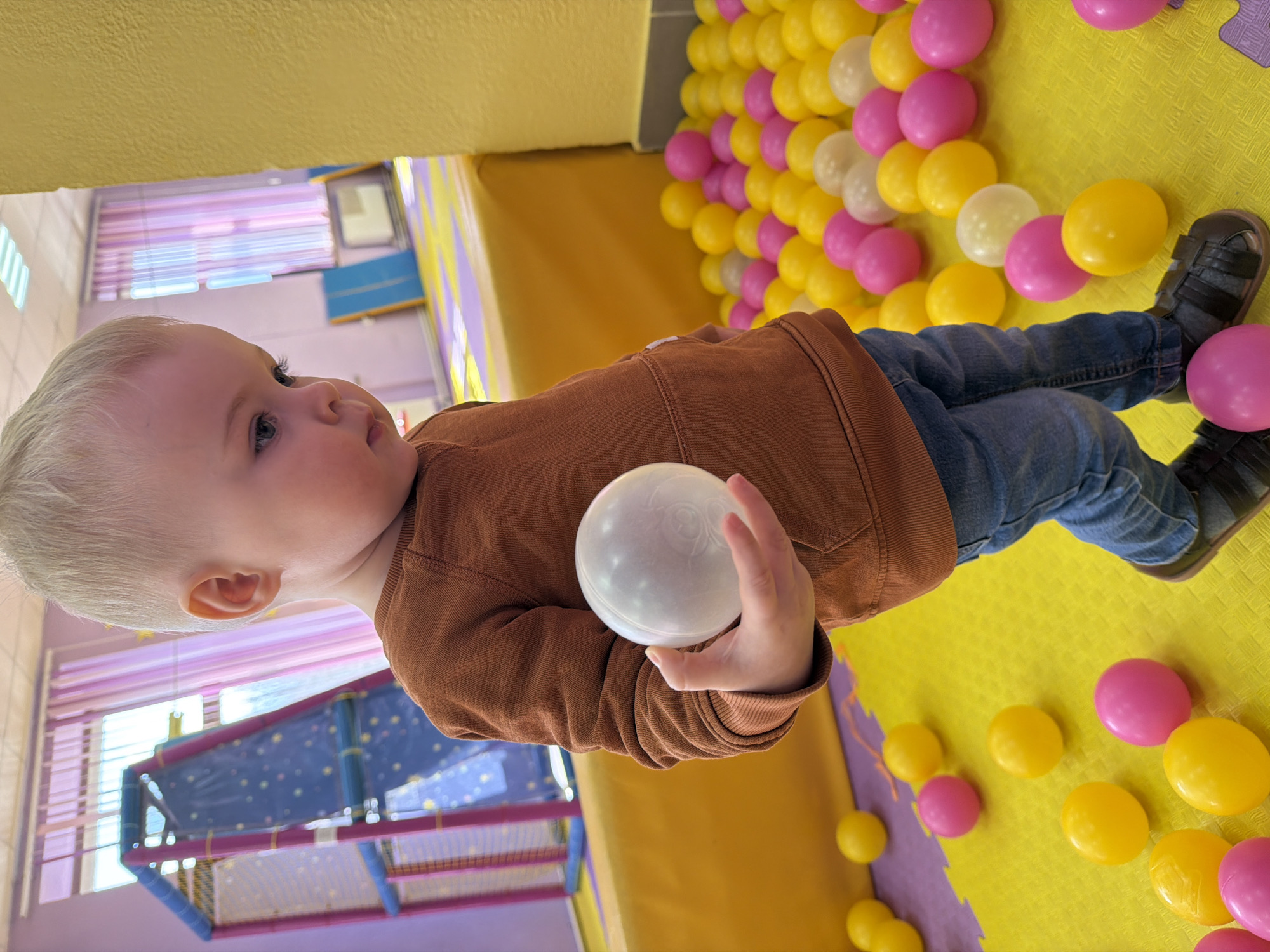 A young child with light blonde hair and fair skin is captured lying on their back in an indoor play area, surrounded by plastic balls. The child is wearing a brown long-sleeved top, blue jeans, and dark sandals. They are holding a translucent white plastic ball in their right hand, looking intently towards the left of the frame.
The setting is a vibrant children's play zone, characterized by a floor covered with yellow and purple interlocking soft mats. Numerous yellow, pink, and white/clear plastic balls are scattered across the floor, indicative of a ball pit. In the background, parts of a multi-level play structure with blue and purple padded frames and protective netting are visible. A window with light purple horizontal blinds and fluorescent ceiling lights can also be seen, suggesting an indoor daytime environment. The child's expression appears calm and curious. This scene likely takes place at a children's entertainment center, potentially located in Votkinsk, Russia. No visible text is present in the image.