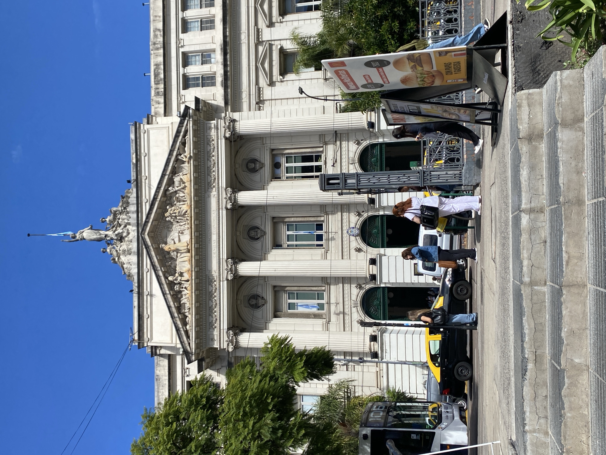 This image depicts a sunny urban street scene in front of the Facultad de Ciencias Económicas (Faculty of Economic Sciences) of the University of Buenos Aires, located on Avenida Córdoba in Buenos Aires, Argentina.
The central subject is a grand, multi-story neoclassical building with a light-colored facade. It features a prominent portico with multiple fluted columns supporting a pediment adorned with sculptural reliefs. A large female statue, holding an upright staff or torch, crowns the peak of the pediment. The text "FACULTAD DE CIENCIAS ECONÓMICAS" is visibly inscribed below the pediment. Ground level features several arched green entrances, some displaying the Argentinian national shield. Argentinian flags (light blue and white) are visible hanging inside multiple upper-story windows.
In the foreground, a concrete sidewalk with worn, multi-tiered steps occupies the lower portion of the frame. The midground shows several individuals on the sidewalk. Approximately six people are visible, primarily women. A woman in a white top and pants stands near a dark lamppost on the left. Next to her, a woman in a black top and light blue jeans leans against the lamppost, holding a black shoulder bag. Further right, a woman in dark clothing walks past two prominent white A-frame advertising signs for McDonald's, which display burger images and "Nuevas Ofertas" (New Offers) in Spanish. Another woman in a dark jacket stands near a black-and-yellow taxi.
On the street, a black-and-yellow taxi is either parked or stopped. The front section of a white and grey public bus (route 106) is visible on the far left, and a silver van is partially obscured behind the taxi. Large green trees, possibly Jacarandas, frame the left side of the building, with additional palm trees and greenery visible on the right. The sky is clear and blue with minimal cloud cover.