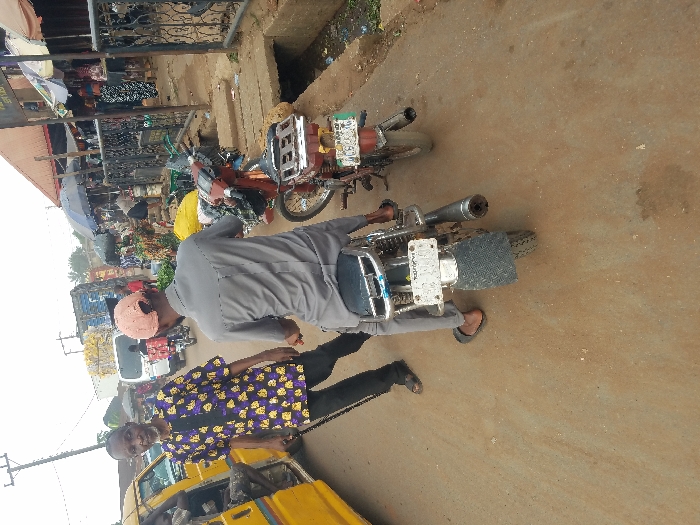 The scene depicts a busy urban street in Nnobi, Nigeria, under bright, overcast daylight.
In the foreground, a light brown dirt road occupies the lower portion of the image. On the left, an older male, dark-complexioned and bearded, wears a purple and yellow patterned long-sleeved shirt over a dark undershirt, dark trousers, and dark sandals. He holds a dark cane in his right hand and faces the camera, positioned next to a yellow commercial minibus with its door open, revealing a passenger's leg inside. To the right of the older male, a second male, dark-complexioned, is seen from the rear, seated on a silver motorcycle. He wears a light grey long-sleeved shirt, light grey trousers, dark flip-flops, and a reversed light pink baseball cap. The motorcycle has a dark blue seat, a white rear license plate, and an exposed exhaust pipe. Adjacent to this motorcycle, on the right side of the road, a second, red-brown colored motorcycle is parked, also facing away from the camera, with a green and white rear license plate and items secured to its rear rack.
The mid-ground reveals a concrete drainage ditch running along the right edge of the road, containing refuse and dark liquid. Behind this ditch, a row of market stalls with dark metal frames and various tarpaulin or umbrella coverings, including a prominent dark blue umbrella, display goods. Yellow sacks are visible near these stalls.
The background shows multiple pedestrians and vehicles, indicating a high level of activity. A white van is visible behind the seated motorcyclist, and further in the distance, a large blue truck is present. Buildings with reddish-brown roofs extend behind the market stalls. A tall utility pole with wires is visible on the far left against the bright sky.