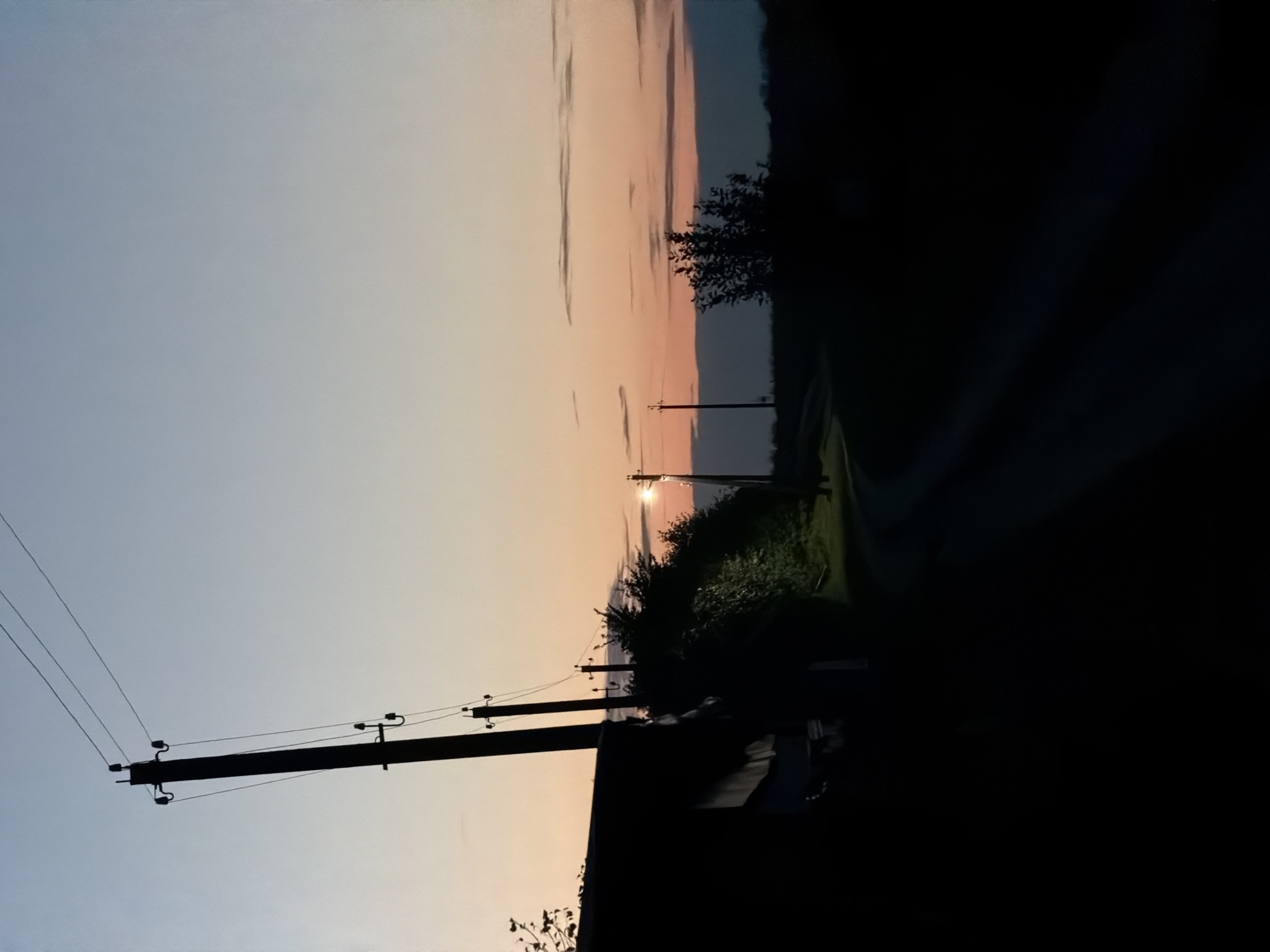 The image depicts a rural scene at dusk, likely in the Валевачскі сельскі Савет, Belarus. In the foreground, a dark silhouette of a building or structure is visible on the left. A dirt road curves into the distance. Several utility poles with wires are scattered across the landscape. The sky displays a gradient of colors, transitioning from a pale blue at the top to shades of orange and pink near the horizon, indicating sunset. A faint light source, possibly a distant streetlamp or the setting sun, is visible on the right side of the image. Trees and bushes are silhouetted against the fading light. The overall atmosphere is one of tranquility and the end of the day in a rural setting.