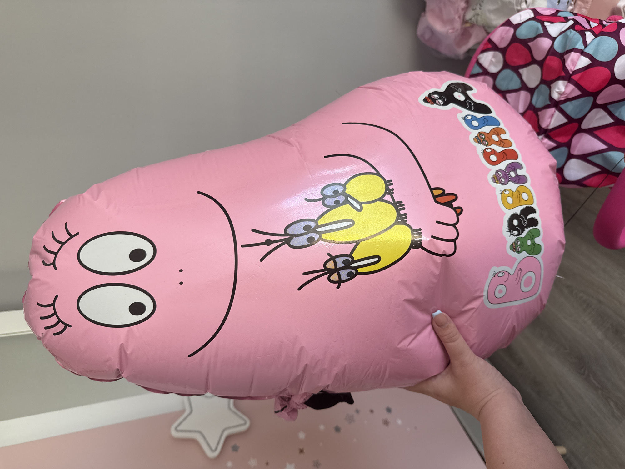 A person's left hand, with pale skin and light blue-painted fingernails, holds a large, deflated pink balloon featuring cartoon characters. The balloon is shaped like an elongated blob with a face at the top, consisting of two large oval eyes with black pupils, simple eyelashes, and a wide, smiling mouth. Below the face, there are two yellow cartoon birds with big eyes and beaks. A banner-like design runs along the bottom of the balloon, displaying the word "BARBAPAPA" in colorful letters, with various characters from the Barbapapa franchise interspersed. The background shows a pale pink wall with a white star decal and a grey wall. In the lower right, parts of a pink, purple, and blue patterned bag are visible. The flooring appears to be light grey wood laminate. There is no discernible information in the image to suggest the location is Burgas, Bulgaria.