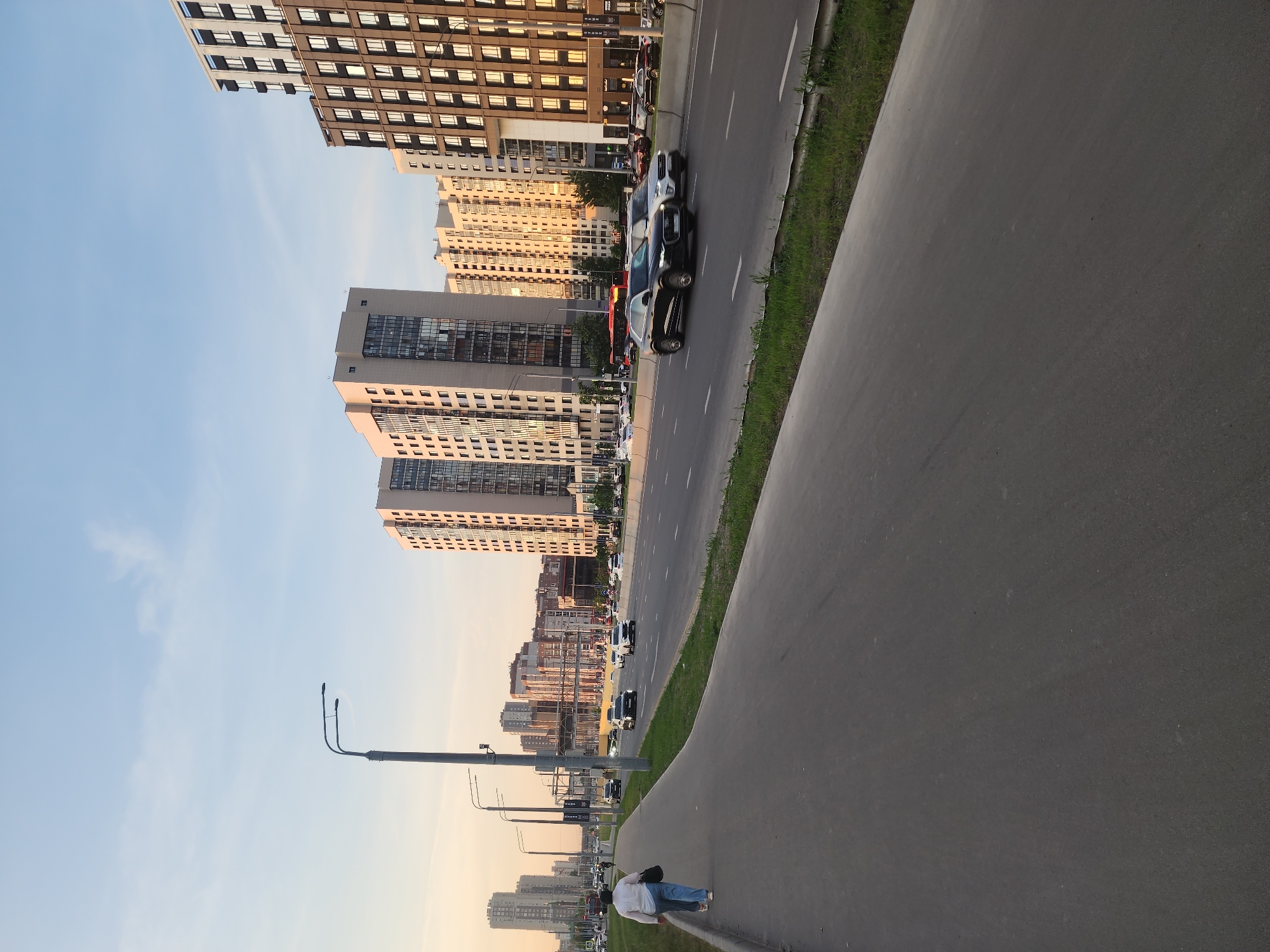 The image depicts a street scene in Kazan, Russia. Tall residential buildings line one side of the street, with a multi-lane road running between the buildings and a wide pedestrian sidewalk. A person wearing a white shirt and blue jeans is walking along the sidewalk. Cars are traveling on the road. Streetlights are visible along the sidewalk. The sky is light blue.