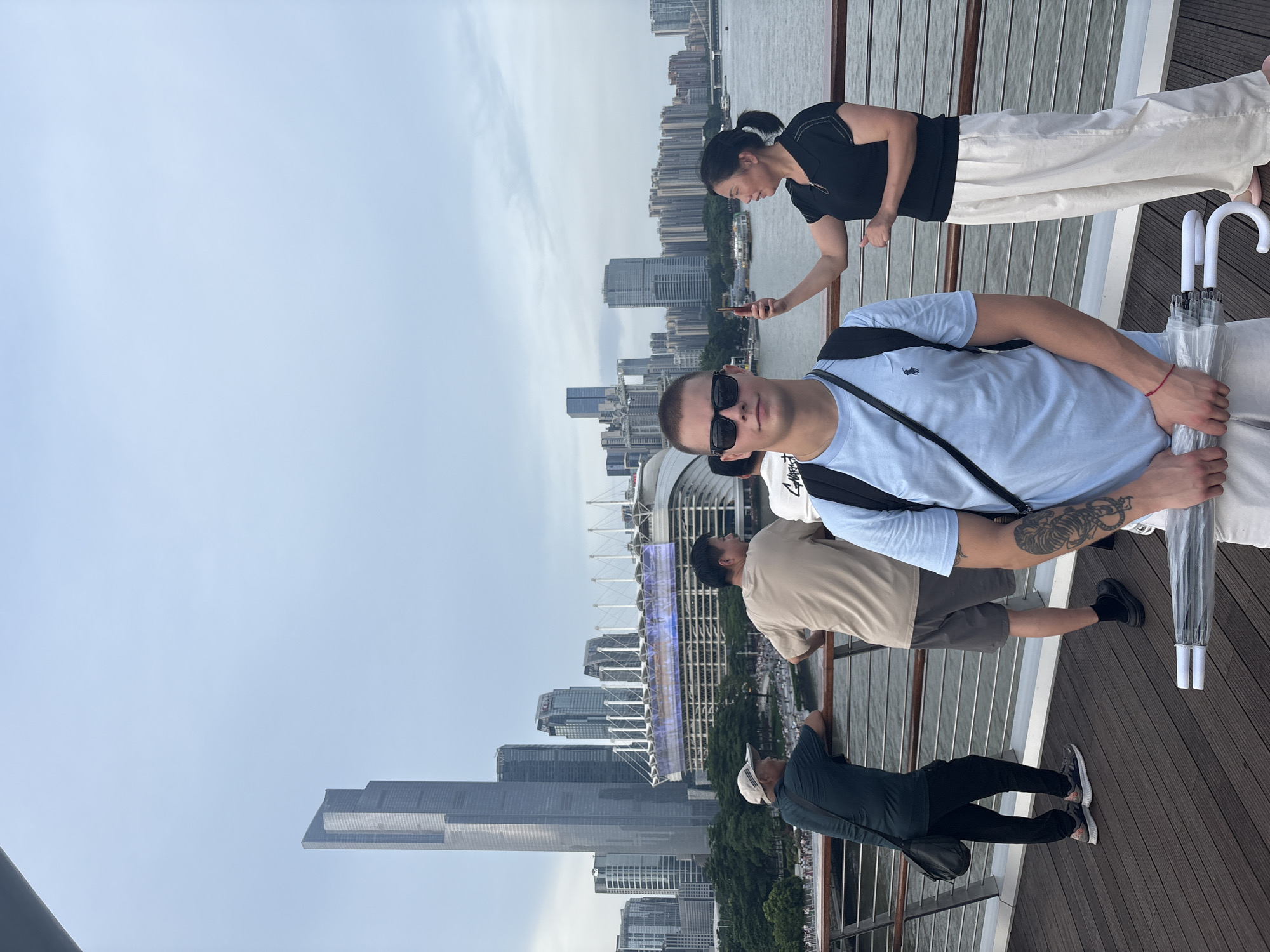 This is a medium shot of a group of people on a wooden boardwalk with a city skyline in the background. In the foreground, a young man with sunglasses and a light blue t-shirt stands looking at the camera, holding a clear umbrella. Behind him, a woman in a black shirt and white pants is taking a photo with her phone. Further back, two men are leaning on a railing, looking out at the water and the city. The skyline features tall modern buildings, including a prominent skyscraper, and a large stadium with a colorful illuminated facade. The sky is overcast. The location appears to be along a waterfront in Yuexiu District, China.