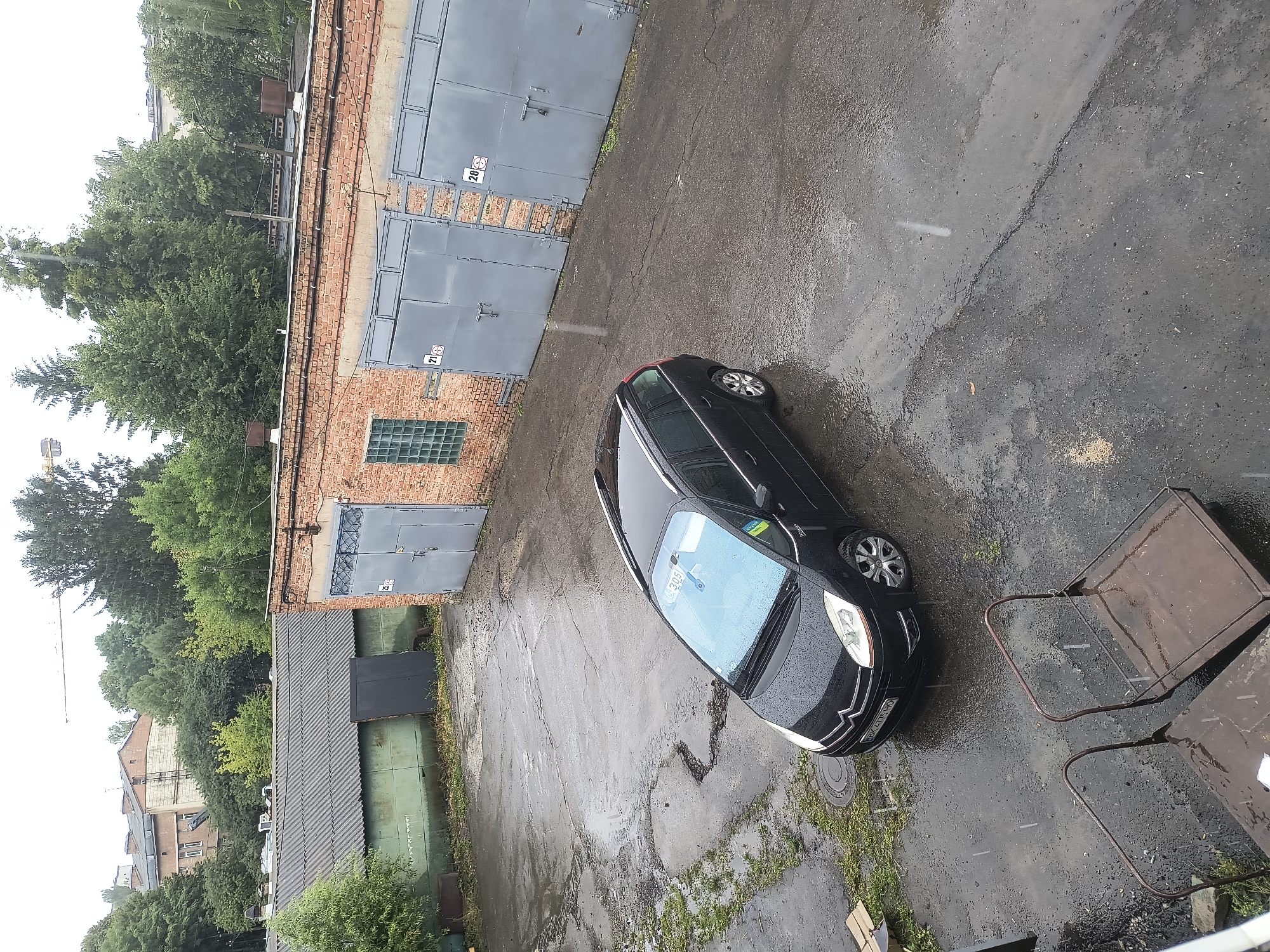The image shows an outdoor scene in the city of Rivne, Ukraine, on a rainy day. The primary focus is a black Citroën C4 Picasso minivan parked on a wet asphalt surface. The minivan is facing towards the left of the frame, with its headlights on. Its windshield is covered in raindrops, and a blue sticker is visible on the upper right corner of the windshield. The license plate of the car reads "BK 4775 HO".
Behind the car, a row of brick garages with grey metal doors is visible. Two of these garage doors are numbered "21" and "20". To the left of the garages, there is a long, grey, corrugated metal building with a dark grey door. The ground is covered in puddles and reflections of the surrounding environment, indicating recent rainfall. Some patches of green grass are growing in the cracked asphalt. In the foreground, two metal carts with wire mesh baskets are present.
The background consists of trees and buildings, including a taller brick building with multiple windows and a construction crane visible above the treeline. The overall atmosphere of the image is overcast and damp, characteristic of a rainy day.