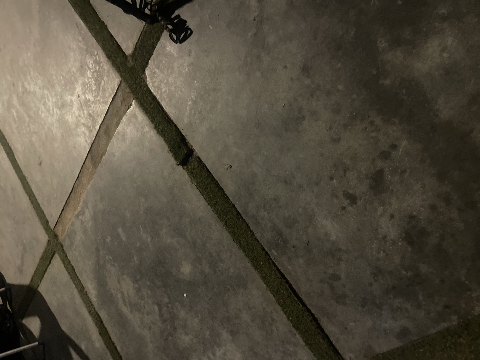 The image shows a concrete patio with artificial turf inlays, laid out in a cross pattern. The concrete is a dark gray color and appears to be slightly wet or stained in places. The artificial turf is a vibrant green and fills the gaps between the concrete slabs. There are some shadows on the patio, suggesting the presence of an overhead light source or perhaps natural light from a low angle. In the upper left corner, part of a dark object, possibly furniture or equipment, is visible, casting a shadow. In the upper right corner, a black object with some tangled cords is present, possibly an electrical cord or a piece of equipment. The scene is outdoors and appears to be nighttime or very early morning due to the lighting conditions. The location is identified as the city of Jimeta, Nigeria.
