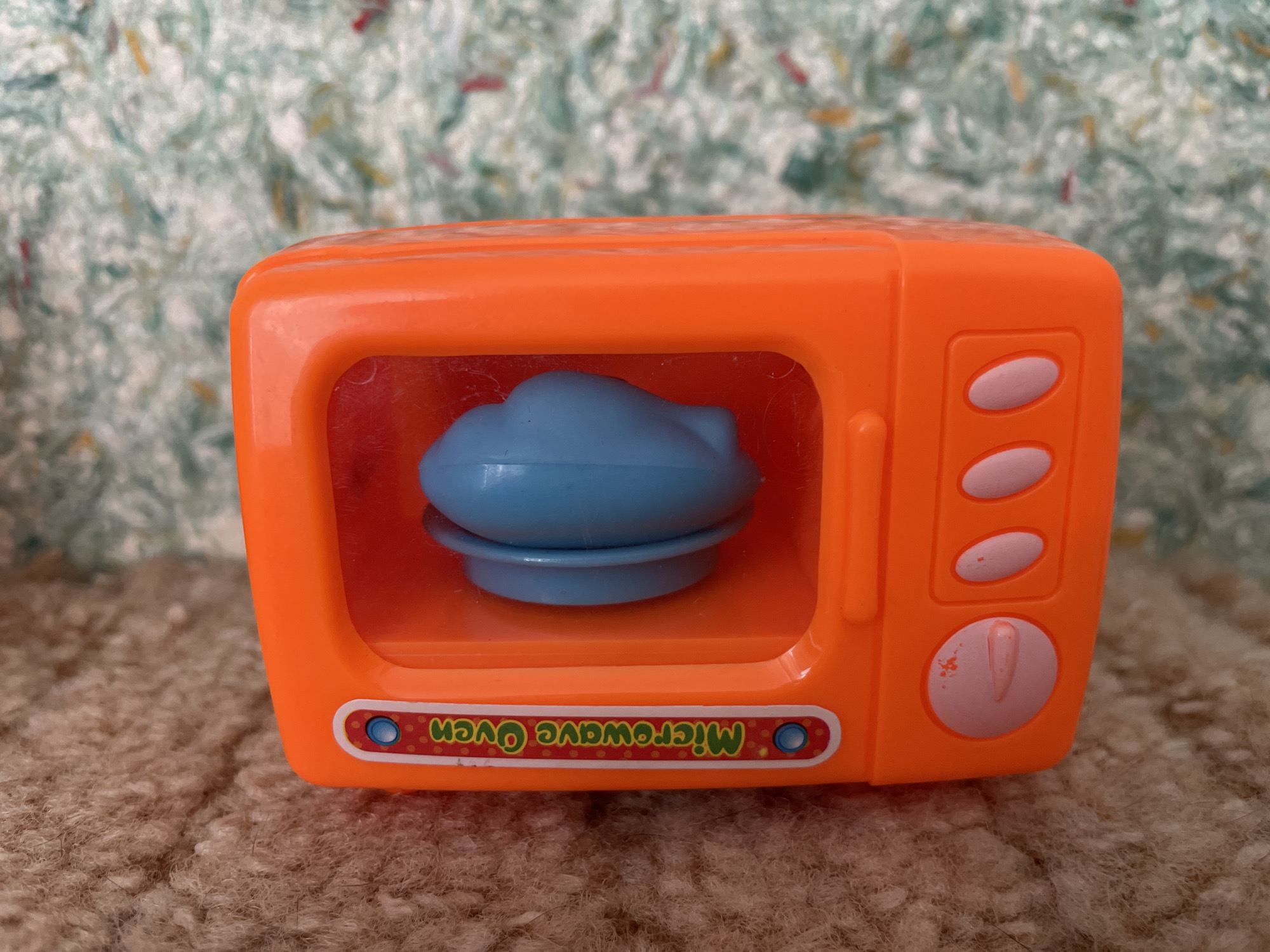A bright orange toy microwave oven is positioned on a textured brown rug. The microwave has a clear plastic door through which a blue toy food item, resembling a cloud or a bun on a plate, is visible. The oven features several pink oval buttons on the right side and a decorative sticker at the bottom with the text "Microwave Oven" written in a colorful, stylized font. The background is a blurry, light-colored fabric with a speckled pattern. There is no indication of the city of Perm, Russia, or any people in the image.