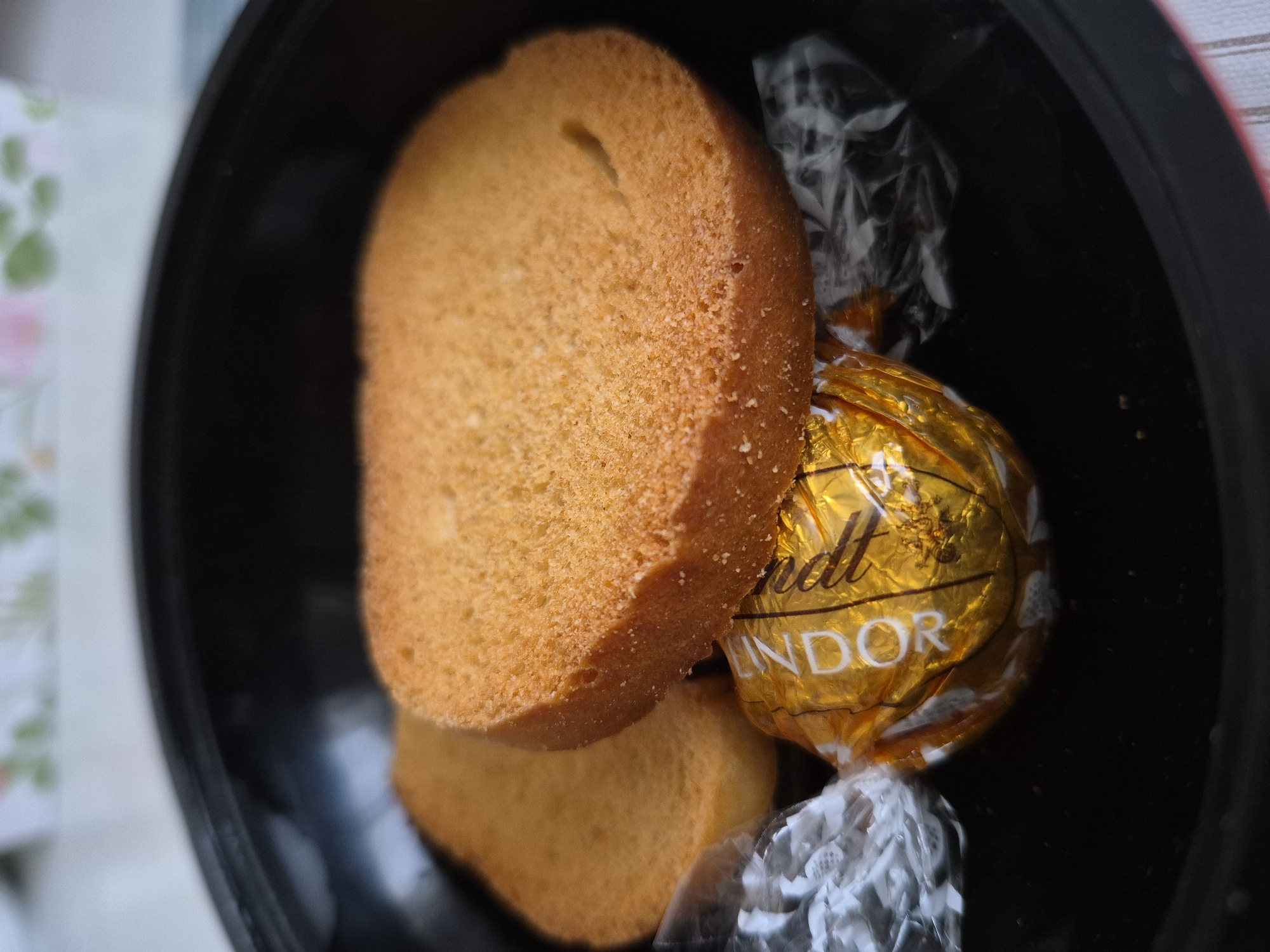 In an overhead close-up shot, two round, golden-brown slices of biscotti rest inside a dark, round bowl. The biscotti are positioned side-by-side, with the right slice slightly overlapping the left. Beneath the biscotti, partially obscured, is a single, shiny, gold-wrapped Lindt chocolate. The chocolate wrapper is slightly crinkled and reflects the light. The bowl appears to be made of a dark material, possibly ceramic or plastic, and has a subtle sheen. The background is blurred, showing hints of a light-colored surface with a floral pattern. The lighting is soft, creating gentle shadows. The scene is static, with no visible action or people.