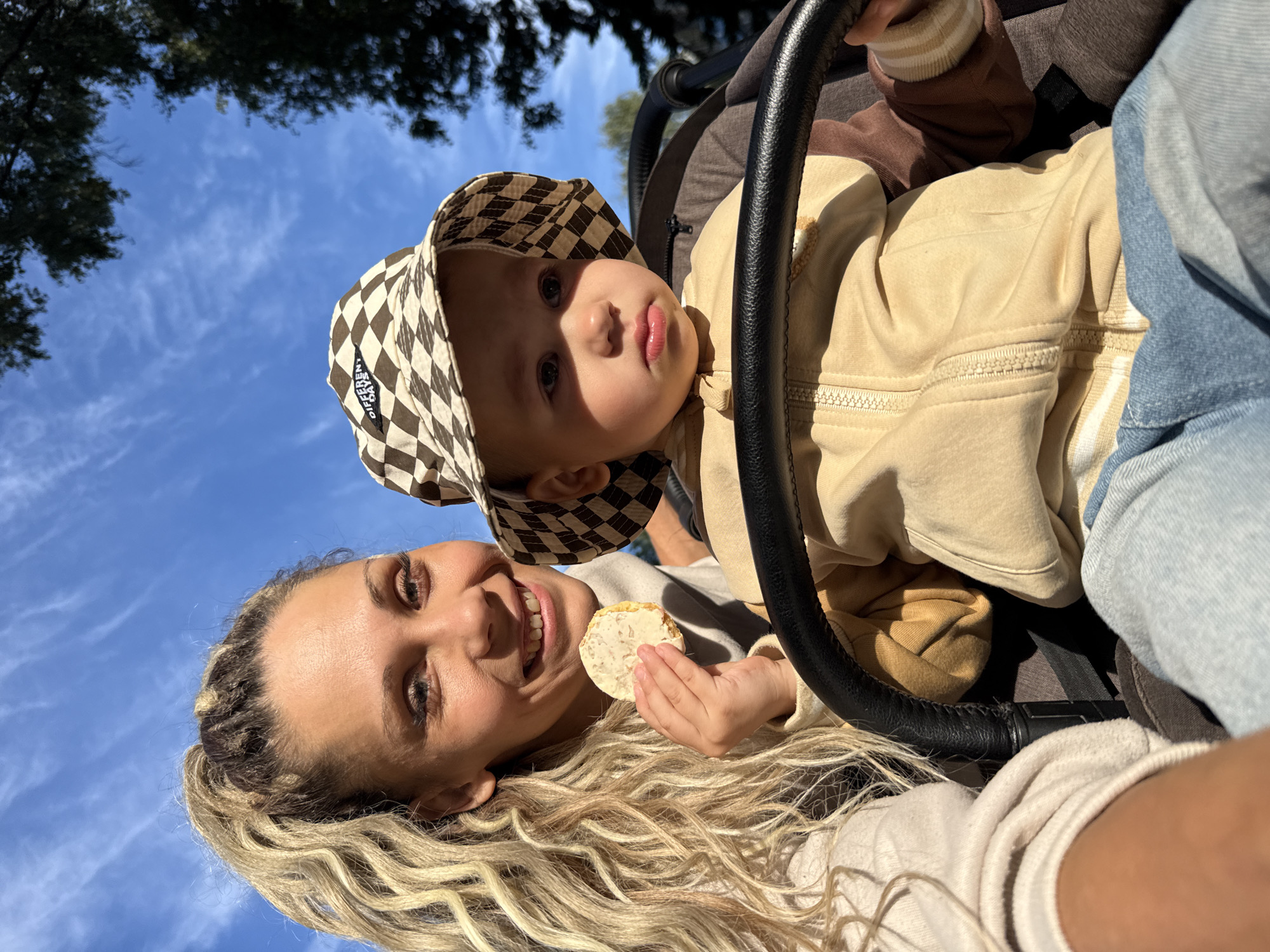 A woman and a young child are in a stroller outdoors. The woman has blonde curly hair pulled into a ponytail and is holding a cookie with a white spread. The child is wearing a bucket hat with a checkered pattern and a tan and brown hooded jacket. The child is seated in the stroller, holding onto the stroller's handle. The background shows a blue sky with white clouds and the silhouette of trees. The scene appears to be in a park or outdoor recreational area.