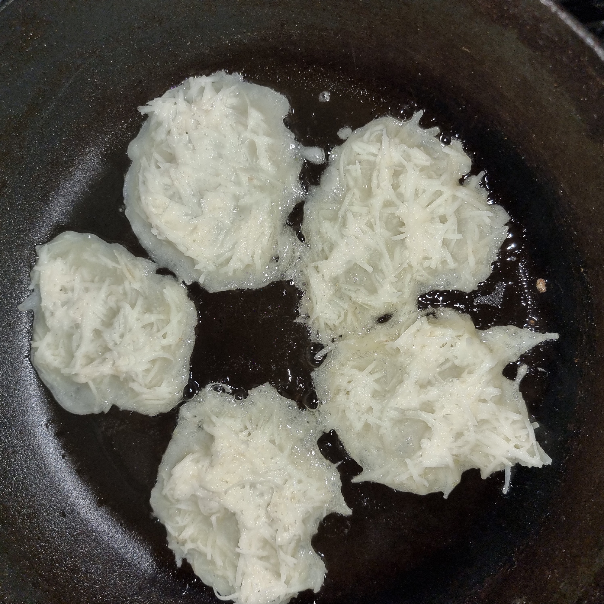 Four potato pancakes are being fried in a dark, cast-iron skillet with a black background. The pancakes are roughly circular and composed of shredded potatoes. They are submerged in hot oil, which is bubbling around them. The oil is a dark, glossy brown, with lighter oil pooling around the edges of the pancakes. The skillet's surface is also dark and slightly textured. The lighting is dim, suggesting an indoor setting, possibly a kitchen. The overall scene depicts the cooking process of potato pancakes. No people are visible. There is no visible text. The location context is Dunnae-myeon, South Korea.
