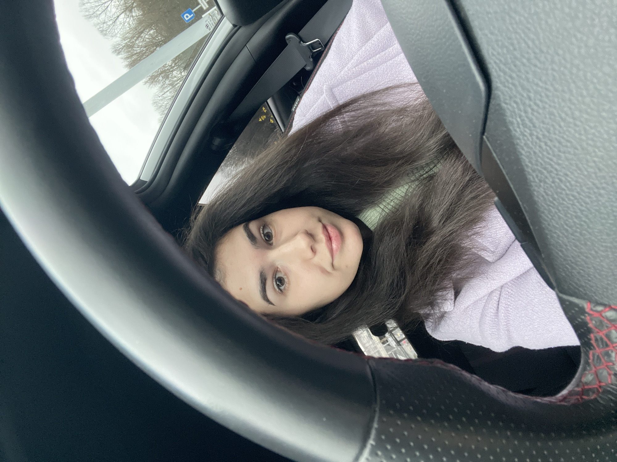 A selfie taken from inside a vehicle, presumably a car, showing a young woman with long, dark hair looking at the camera. The steering wheel is prominently featured in the foreground, partially obscuring the view. Behind her is a passenger seat covered with a light-colored fabric, and a seatbelt is visible. Through the car's windshield, trees and a signpost with a parking symbol are visible. The signpost suggests the location is in a parking area. The photo does not contain sufficient visual information to determine whether it was taken in Sofia, Bulgaria.