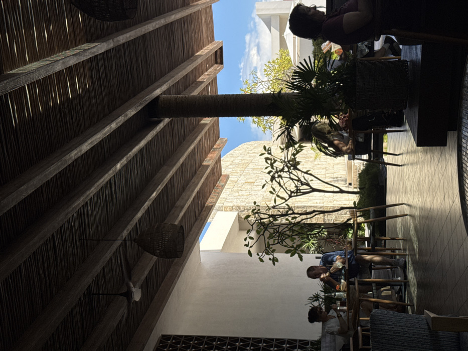 The image displays an outdoor or semi-outdoor dining area located in Marina Maroma, Mexico, under clear daylight conditions. The scene is viewed from a low perspective, focusing on the shaded dining space and elements beyond it.
Overhead, a dark wooden slatted ceiling is supported by parallel wooden beams. A single-blade ceiling fan is visible towards the top left, and a dark, woven, teardrop-shaped pendant lamp hangs centrally. Sunlight filters through gaps in the ceiling, creating bright streaks.
On the left, a smooth white wall features a geometric lattice pattern at its upper section. To the right of the dining area, a large, textured light-colored stone wall, possibly coral, curves gently. Further in the background, under a clear blue sky with sparse white clouds, white building structures are visible, including a distant white canopy or umbrella. A tall, dark, cylindrical pillar, appearing to be wrapped in a natural material like palm fiber, extends from the floor to the ceiling, centrally positioned on the right side of the frame.
Four individuals are present, seated at wooden tables. In the mid-left, a female with dark, pulled-back hair, wearing a light-colored top, is seated, looking down at objects on her table. Opposite her, a male with short, light-colored hair, dressed in a blue short-sleeved shirt, is seated, observed with his hand near his mouth, indicating consumption. A bottle and small dishes are on their table. Further right, partially obscured by a potted plant, a third female with lighter hair is seated at another table. On the far right, a fourth female with dark hair, possibly wet, wearing a dark top, is seated in profile, looking towards the right side of the frame, with her right arm resting on the table.
The floor is covered with light-colored, geometrically patterned tiles. Multiple plants enhance the environment, including two smaller trees with sparse branches in a landscaped bed between the dining area and the stone wall, and a large potted plant with broad leaves in the mid-right foreground. Strong directional sunlight illuminates the background stone wall and patches of the tiled floor, contrasting with the deep shadows in the immediate foreground and within the shaded seating areas.