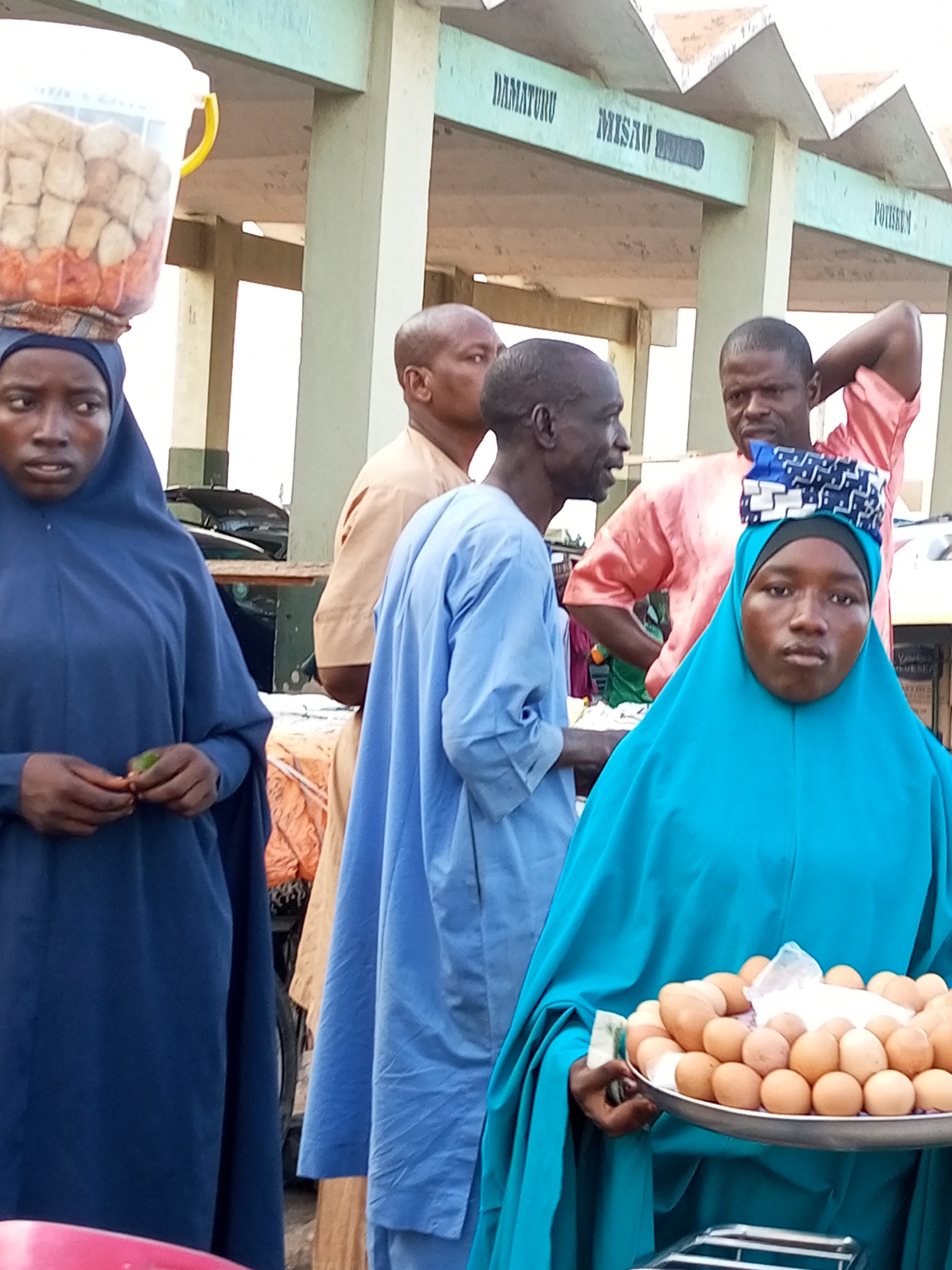 A group of people are standing in what appears to be a market in Gombe, Nigeria. In the foreground, a young woman wearing a blue hijab is holding a tray of eggs. Behind her, another woman in a blue hijab is carrying a bucket of food on her head. Several men in traditional clothing are also present. The background shows a building with signs indicating "DAMATURU," "MISAU," and "POTISKUM," which are likely locations in the region. The overall scene depicts a bustling marketplace atmosphere.