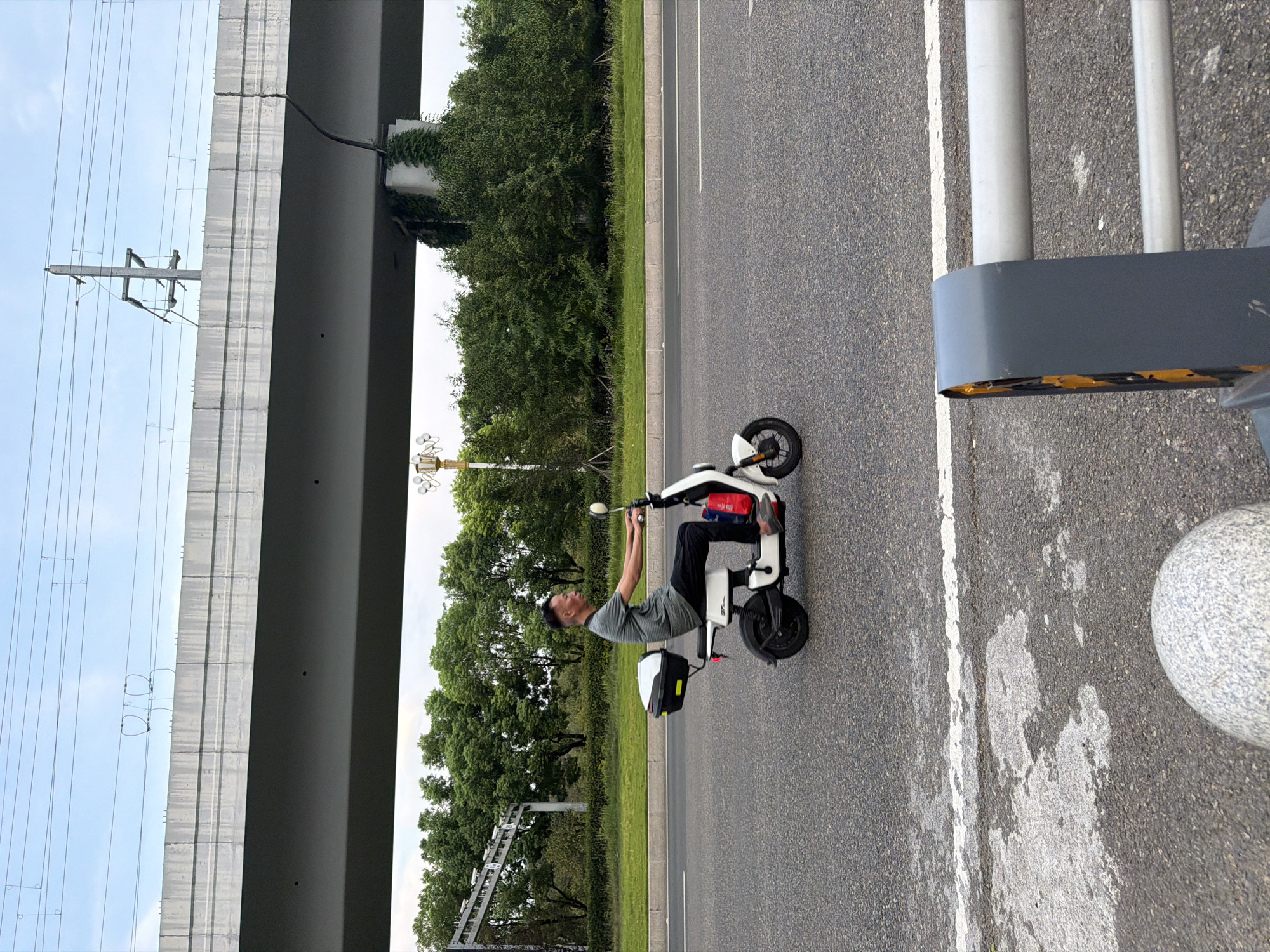 A man rides a white electric scooter down a paved road in Jiangjin, China. He is wearing a grey, striped t-shirt and black pants. The scooter has a black and white storage box on the back and a red container on the side. In the background, there is a large elevated concrete structure, possibly an overpass, with power lines above it. Trees and vegetation line the side of the road. The sky is cloudy.