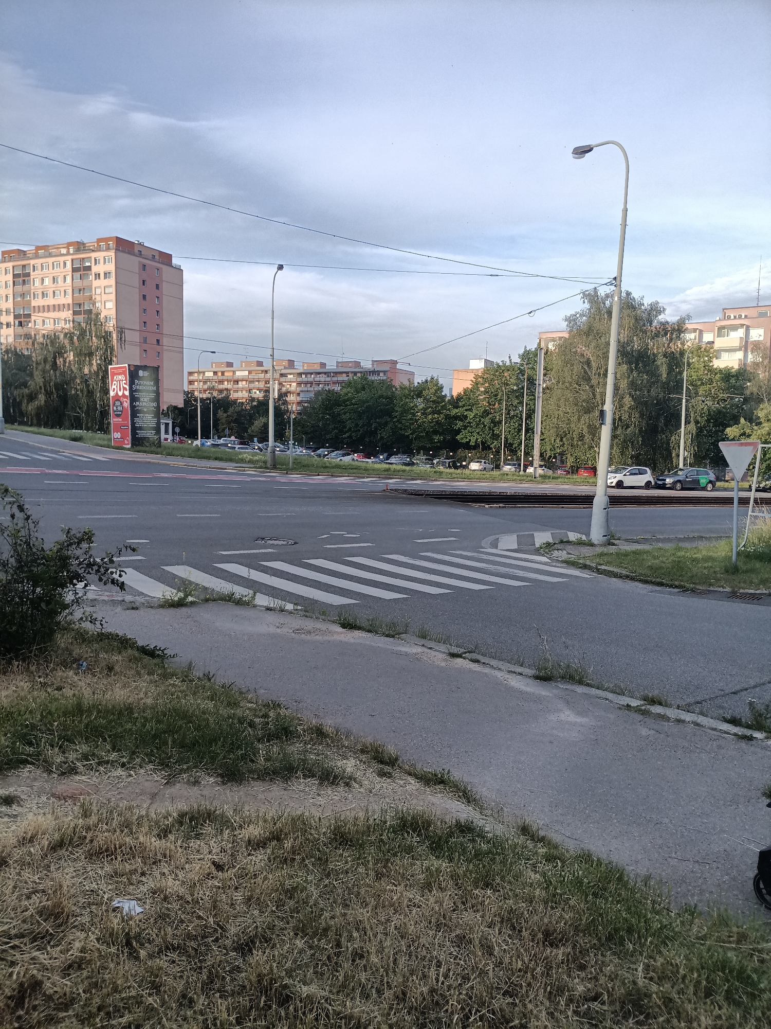 The image captures an outdoor scene in Prague, Czechia, likely during twilight or overcast conditions. The foreground features a dry, grassy verge adjacent to an asphalt road with a pedestrian crossing marked by white stripes. The road curves slightly to the right. Beyond the crossing, tram tracks are visible. In the middle ground, several parked cars are situated in front of a row of trees. The background is dominated by apartment buildings with light-colored facades and numerous windows, characteristic of residential areas in Prague. A tall street lamp stands prominently on the right side of the image. Overhead, power lines stretch across the sky, which is partly cloudy. A large advertisement panel for "KINO BUS" is visible on the left, suggesting a cultural venue nearby. No people are clearly depicted in the image.