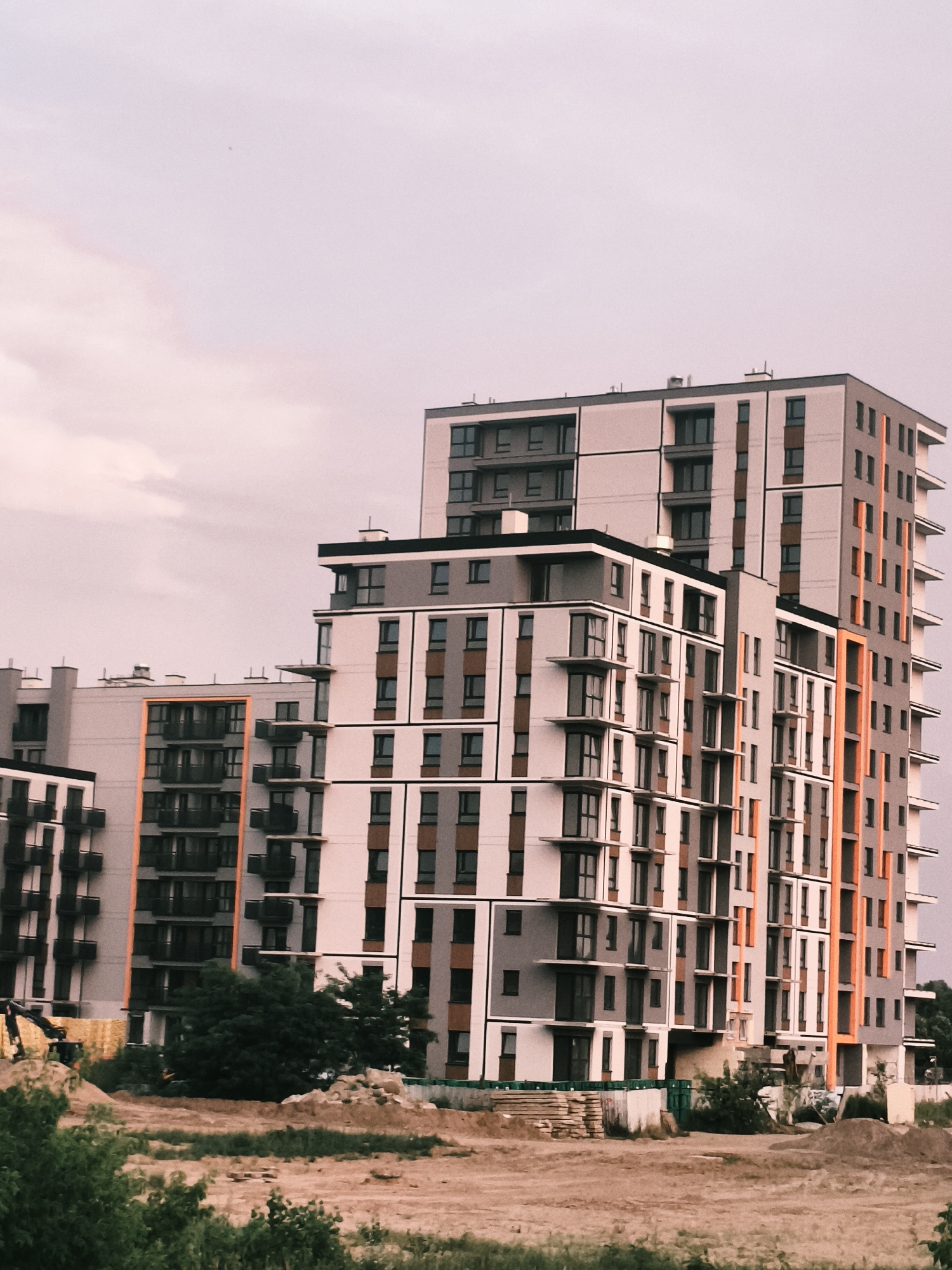 This image depicts a modern residential building complex under construction in Nowy Dwór Mazowiecki, Poland. The primary subjects are the multi-story buildings, characterized by their contemporary architectural design featuring a combination of light grey, white, and orange facades, with numerous windows and balconies. In the foreground, there is an area of disturbed earth, indicative of ongoing construction, with piles of soil and what appear to be construction materials such as wooden planks and possibly fencing. A yellow excavator is visible to the left, suggesting active construction work. Green trees and foliage frame the bottom and sides of the image, contrasting with the urban development.
The sky is a soft, muted pinkish-grey, suggesting either early morning or late evening light, possibly during twilight. The weather appears to be overcast, with subtle cloud formations visible. There are no people visible in the frame, and the focus is on the architecture and the construction site. The overall mood is one of development and progress, with the modern buildings standing starkly against the natural elements and the signs of construction. No visible text is present in the image.
