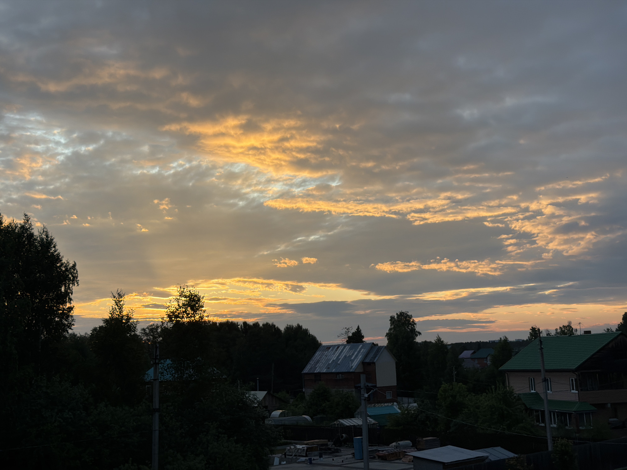 The image depicts a rural or suburban landscape at sunset, likely in Chaikovsky, Russia, as suggested by the presence of dacha-style houses and dense tree cover common in the region. The sky is dramatic with a mixture of dark grey and orange clouds, indicating a late evening or early morning light. Sunbeams are visible breaking through the clouds, illuminating portions of the sky with a golden hue. In the foreground, silhouetted trees and bushes dominate the lower part of the frame, obscuring some of the details of the settlement. Several buildings are visible, including houses with pitched roofs, some covered in corrugated metal, and others with what appear to be green shingles. The architecture suggests a residential area, possibly a dacha community, characterized by individual houses surrounded by vegetation. There are no visible people or active events taking place. The overall impression is one of quietude and the natural beauty of a twilight sky over a rural dwelling area.