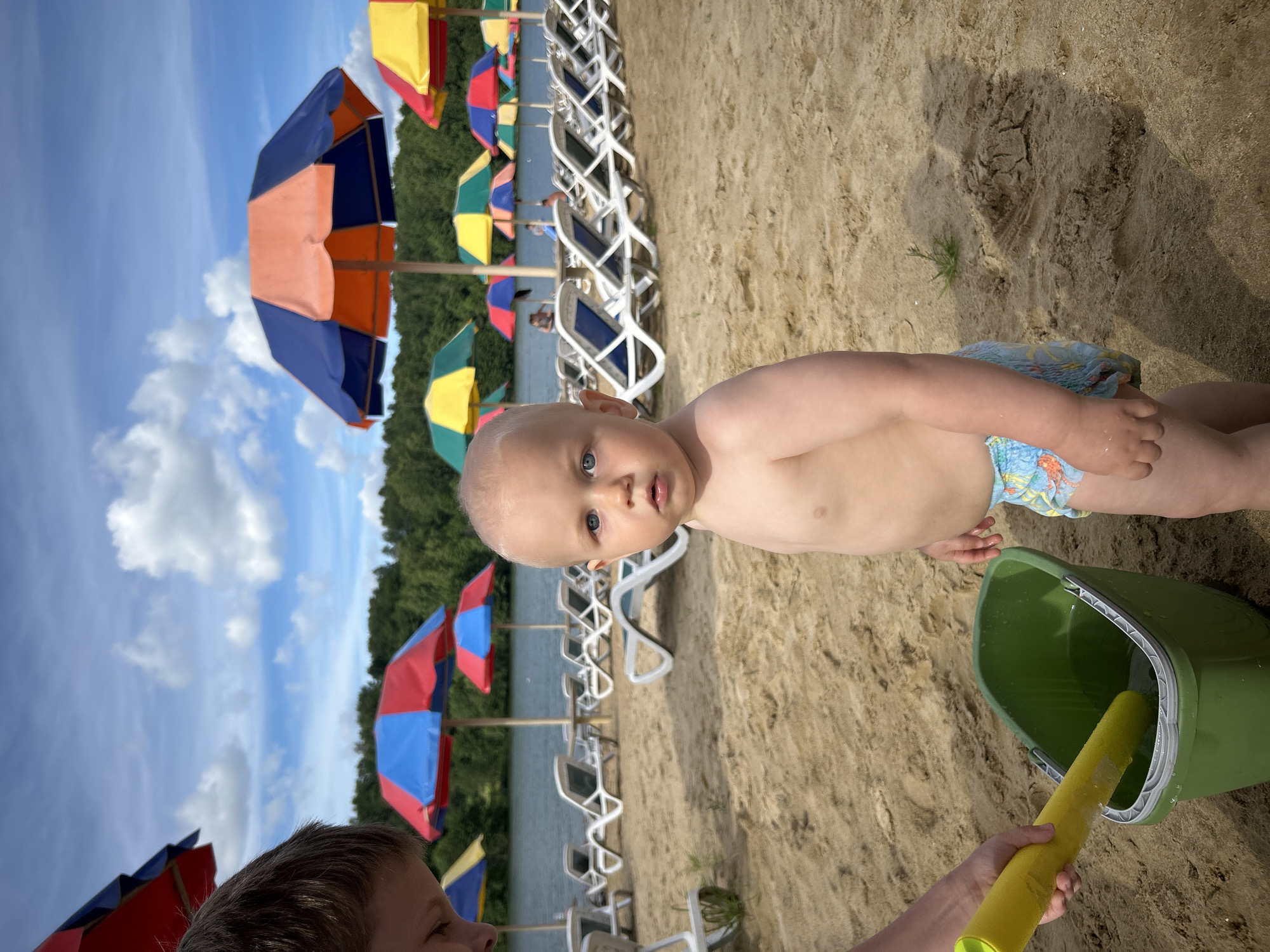 A young child, likely a toddler, stands on a sandy beach in the foreground of the image. The child has fair skin and light hair, with bright blue eyes looking directly at the camera. The child is wearing a diaper with a blue and orange pattern. To the left of the child, a person's arm, presumably belonging to an older child or adult, is visible, holding a yellow plastic shovel and a green bucket filled with water. The background of the image depicts a body of water, likely a lake, with a line of lounge chairs and colorful beach umbrellas visible behind it. Beyond the lake, a dense line of green trees forms the horizon under a partly cloudy sky. The overall scene suggests a recreational area, possibly a public beach within the city of Yubileyny, Russia.