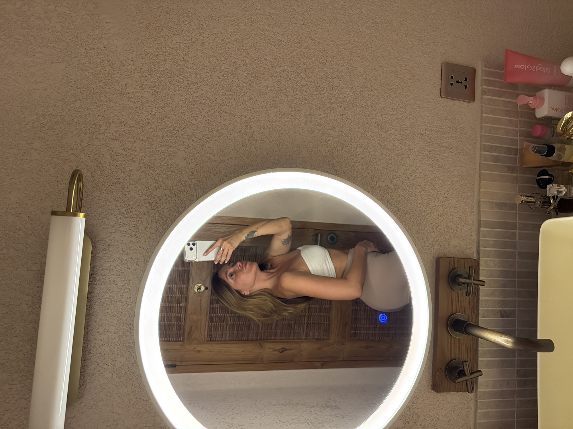 This media file shows a woman taking a mirror selfie in a bathroom, presented rotated 90 degrees clockwise. The scene, if viewed upright, reveals a modern bathroom setting. In the center is a large, circular mirror with an illuminated LED light strip around its circumference. The woman, reflected in the mirror, appears lying horizontally due to the image's rotation. She has long brown hair and wears a white strapless top and light-colored bottoms. She holds a white smartphone with her left hand to take the picture, and a tattoo is visible on her upper left arm.
The background wall is light brown with a textured, stucco-like finish. To the left of the mirror (when viewed upright), there is a tall, vertical light fixture with a white diffuser and gold-colored accents. The reflection in the mirror behind the woman shows a wooden surface, possibly a door or cabinet, featuring a woven or rattan pattern, along with a doorknob. A small blue circular light, likely an indicator or control for the mirror, glows in the reflection.
To the right of the mirror, a multi-socket power outlet is visible on the textured wall. Below it, a small shelf holds various toiletries, including a pink tube with the upside-down text "Glow2Glow." Further right, a contemporary faucet system with a central lever and two cross-shaped handles is mounted on a wooden block against a light-colored, horizontally tiled backsplash. The artificial lighting suggests it's indoors, possibly evening. The overall aesthetic implies a stylish and potentially resort-like environment in Mengwi, Indonesia.