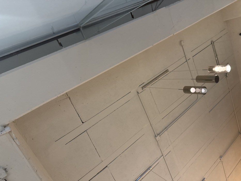 A low-angle shot captures a textured, white ceiling with visible grid patterns. A modern pendant light fixture with three cylindrical shades hangs from the ceiling, casting a soft glow. The architecture suggests an industrial or commercial space. Part of a skylight is visible at the upper left of the frame.