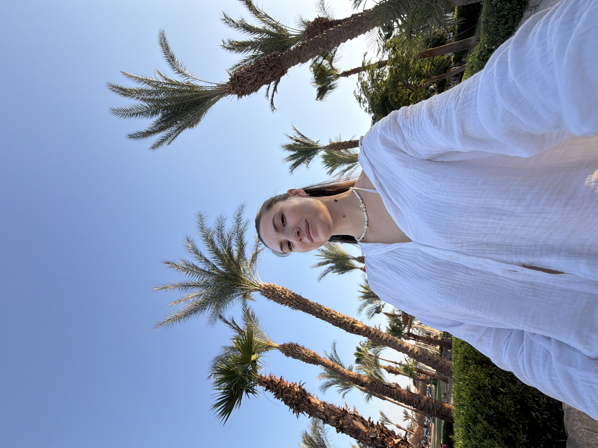 A woman is taking a selfie outdoors in a sunny location, possibly Sharm El Sheikh, Egypt, indicated by the presence of numerous palm trees under a clear blue sky. She is wearing a white, crinkled long-sleeved shirt over a white bikini top and a necklace with small white beads. Her hair is dark and pulled back, and she has a slight smile. The background features a dense arrangement of palm trees of varying heights, some with rough, textured trunks, and lush green bushes. In the distance, there are hints of resort buildings with thatched roofs, suggesting a vacation setting. The lighting indicates it is likely late afternoon or early morning, with soft sunlight illuminating the scene and casting shadows. The image is captured from a low angle, emphasizing the height of the palm trees and the expanse of the sky.