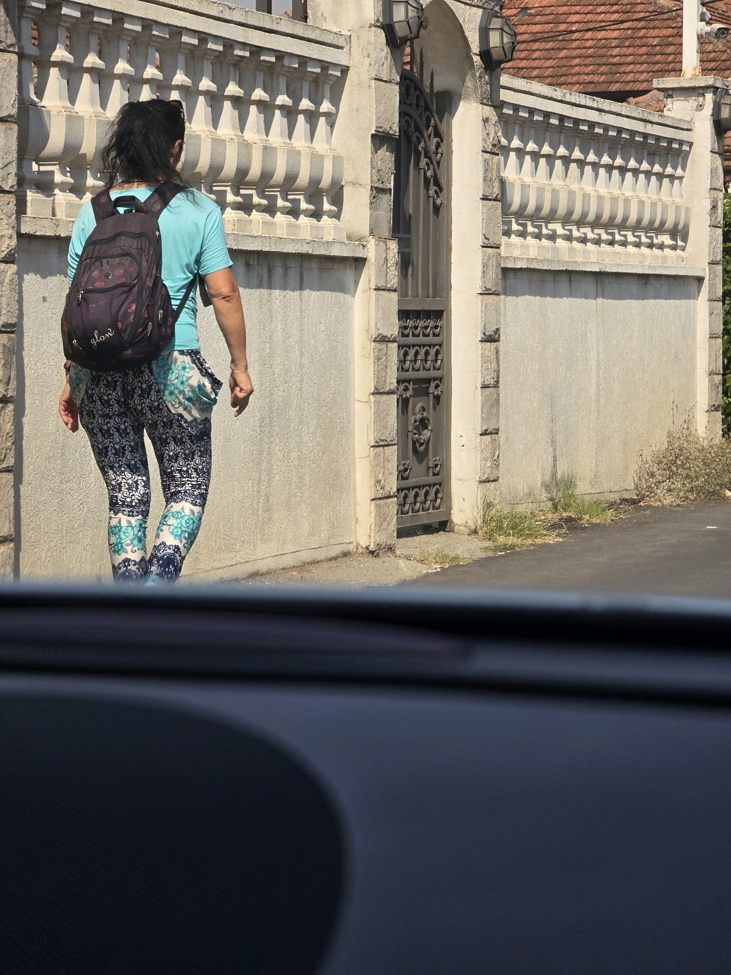 The image shows a woman from the back, wearing a teal t-shirt and patterned leggings, with a dark backpack. She is walking along a sidewalk next to a concrete wall with decorative architectural elements and a metal gate. The scene appears to be in a city, with a road in the foreground and buildings and a red-tiled roof visible in the background. There is some dry vegetation growing near the wall. The overall impression is of a sunny day in an urban environment. While the location is stated to be Podgorica, Montenegro, there are no specific identifying landmarks or contextual clues within the image itself to confirm this.