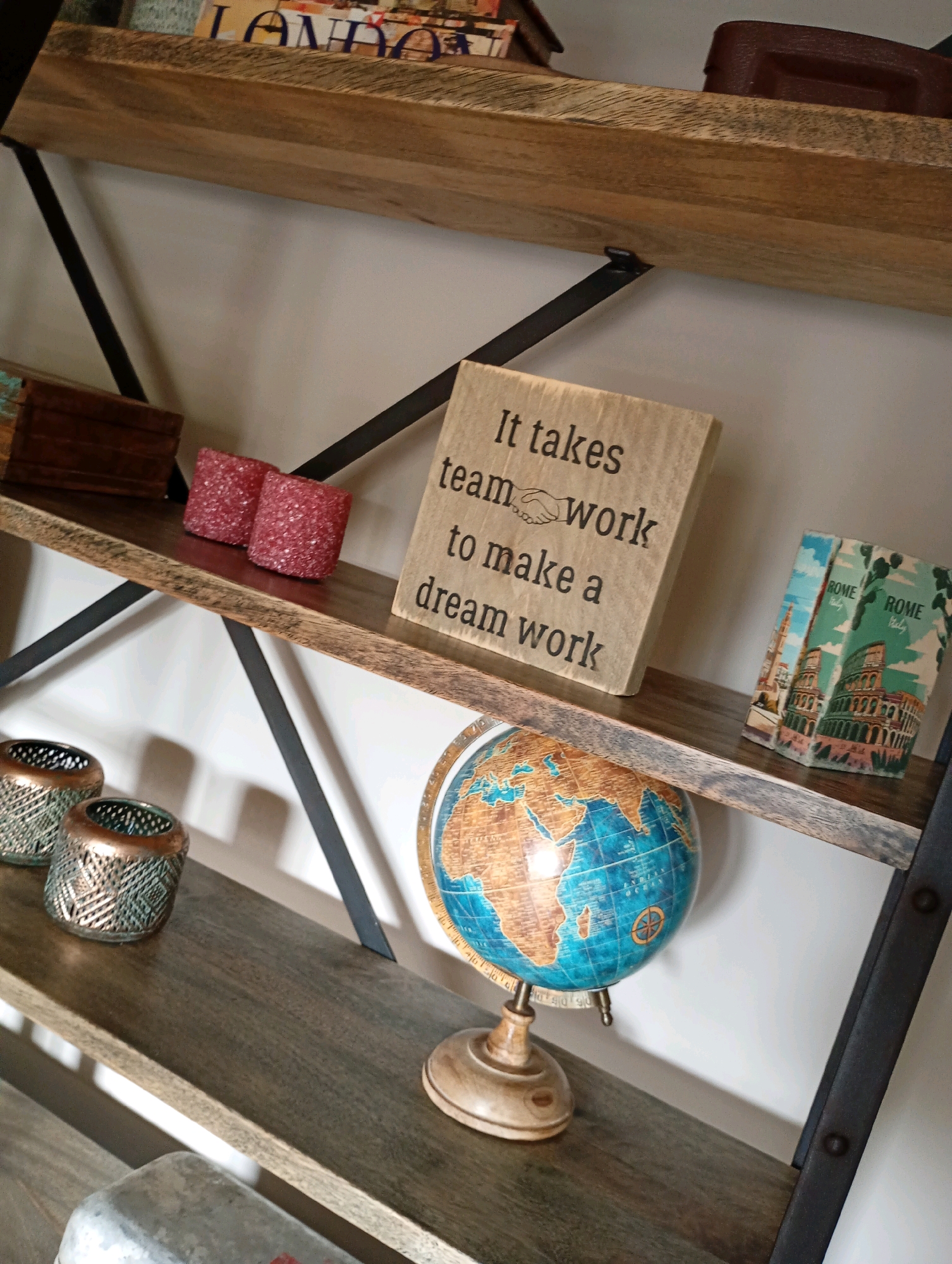 The image shows a wooden bookshelf with several decorative items. On the top shelf, there are books, one with the word "LONDON" visible. To the right of the books, there is a brown leather-bound item. The middle shelf features two pink, glittery candles, a wooden block with text that reads "It takes team work to make a dream work" accompanied by an illustration of two hands shaking, and two small books with images of Rome on their covers. The bottom shelf displays a globe on a stand, and two metallic candle holders. The bookshelf is made of wood with black metal supports. The background is a plain white wall. The image does not provide any information about the city of Bad Sachsa, Germany.