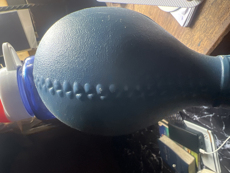 The scene is depicted indoors in the city of Egan, Nigeria, likely on a desk or table surface. No individuals are present.
The primary subject is a large, dark blue, textured, bulbous object dominating the foreground and center. It features a prominent vertical line of raised, irregularly shaped bumps or nodules, reminiscent of a stitched seam or a grip pattern. The surface exhibits a finely stippled texture.
Behind and partially obscured by this blue object, a transparent blue bottle with a white and red flip-top cap is visible. To the left background, resting on a black and white marble-patterned surface, are several objects: a dark blue book, a book with a light-colored spine, and a black rectangular electronic device (possibly a power bank or smartphone) connected to a white charging cable. The cable extends downwards out of the frame. To the right background, a stack of white papers or cards rests on a light brown wooden surface. The lighting suggests an indoor environment with ambient illumination.
