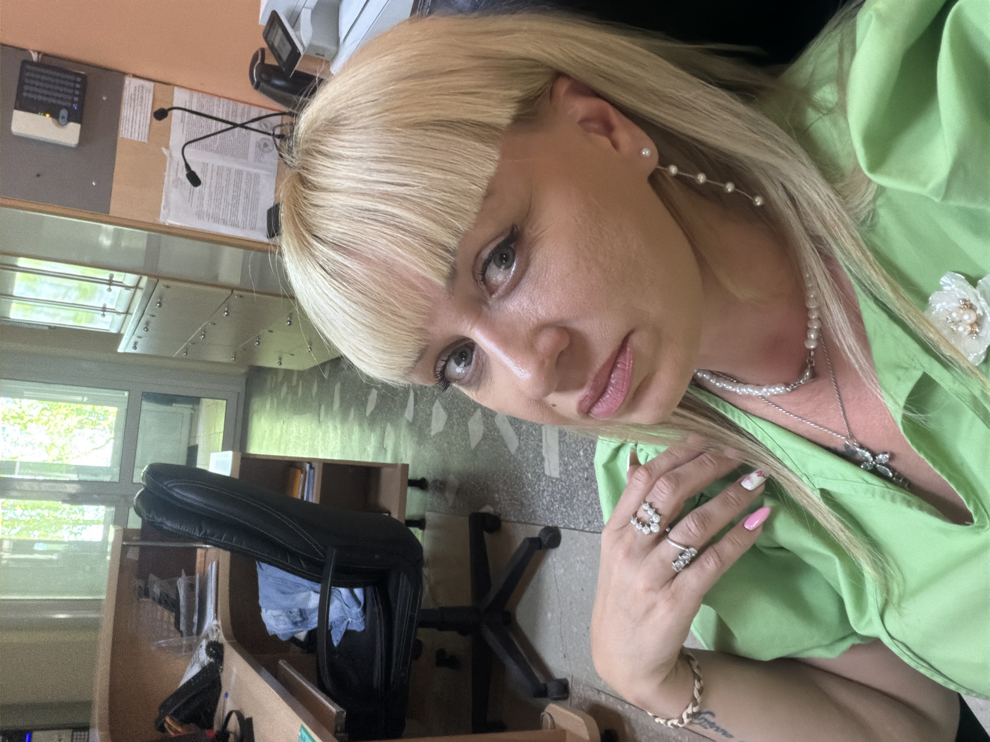 The image presents a woman with blonde hair and blunt bangs, wearing pearl earrings and a matching necklace, as well as rings on her fingers. She is dressed in a light green blouse. The setting appears to be an office or administrative space, with a desk, a black bag, and a printer visible in the background. A window reveals some greenery outside, potentially indicating an urban environment. There's no specific information suggesting the exact location of this image is Chaikovsky, Russia.