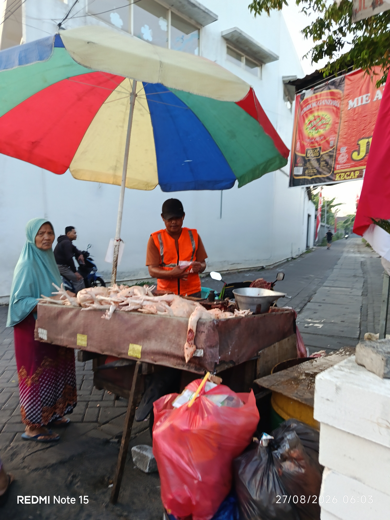 In Keboansikep, Indonesia, an outdoor scene depicts a street-side market stall under a colorful, multi-panel umbrella. A woman wearing a teal hijab and a patterned skirt stands to the left, facing forward. To her right, a man in an orange safety vest and a black cap is arranging items on a wooden cart. The cart is laden with raw chickens, some whole and others appearing to be cut. A metal basin filled with what looks like meat sits on the cart, alongside other debris and a red plastic bag overflowing with trash. In the background, a man on a blue motorcycle is visible, and a street stretches into the distance, lined with buildings and trees. Banners advertising "Mie Ayam" and "Kecap" are hung on a wall. The timestamp "27/08/2026 06:03" and "REDMI Note 15" are visible in the lower portion of the image.