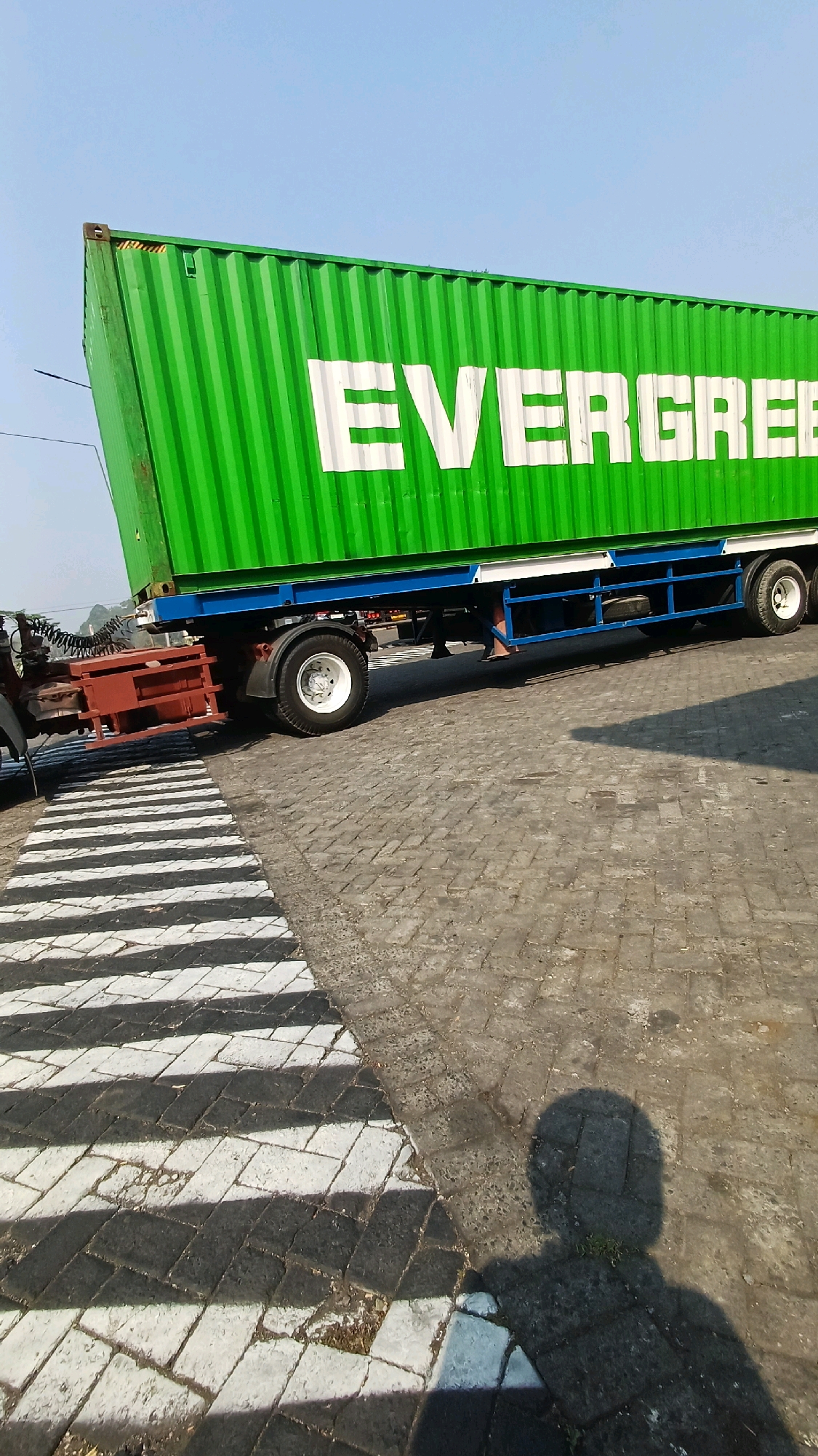 In Kranggan, Indonesia, a green shipping container with the white text "EVERGREEN" emblazoned on its side is loaded onto a truck. The container is positioned over the rear axles of the truck, which has a blue chassis and red cargo box behind the cab. The truck is situated on a paved area with a zebra crossing marked with white and black stripes. A shadow of a person is cast on the ground in the lower right portion of the image, indicating the presence of a photographer. The sky is clear and blue.