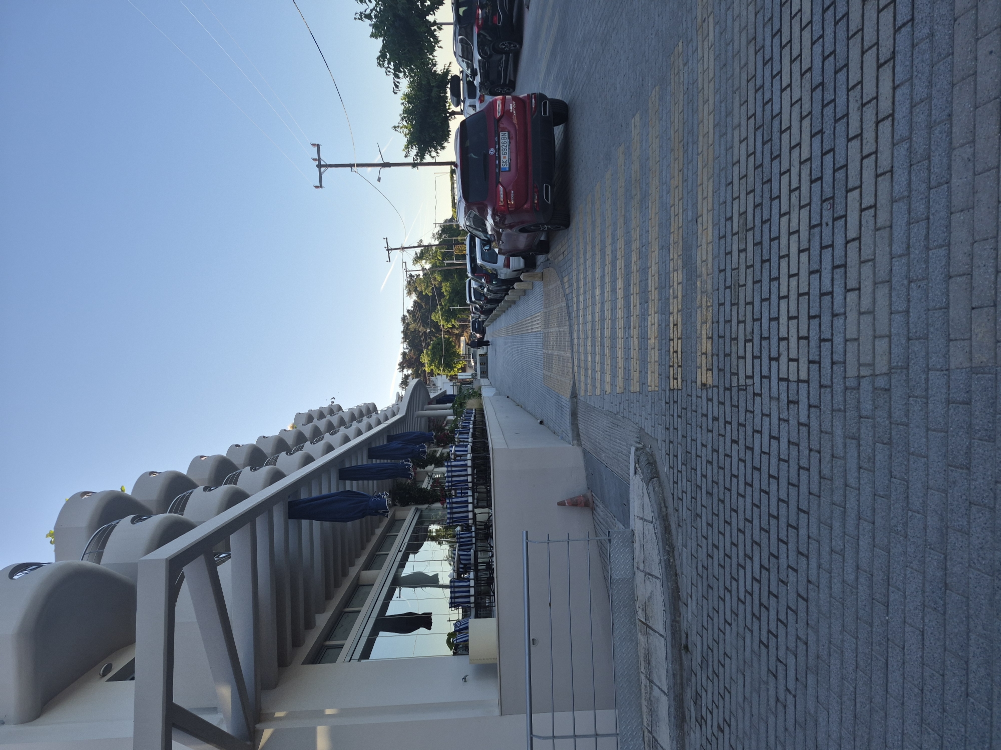 The image shows a street scene in Νέα Καλλικράτεια, Greece, with a modern building on the left and parked cars on the right. The building features a contemporary design with curved balconies and large glass windows, suggesting it might be a hotel or apartments. In front of the building, there is an outdoor seating area with tables and chairs, possibly for a restaurant or cafe, and several closed blue umbrellas are visible. The street is paved with grey interlocking bricks, and a pedestrian crossing with yellow and grey stripes is in the foreground. A row of cars is parked along the right side of the street, including a red SUV with a license plate that reads "SK 6926 BN". A single person is walking away from the camera down the sidewalk. Utility poles with wires are visible against a clear blue sky. The overall environment suggests a pleasant, possibly seaside, location during daylight hours.