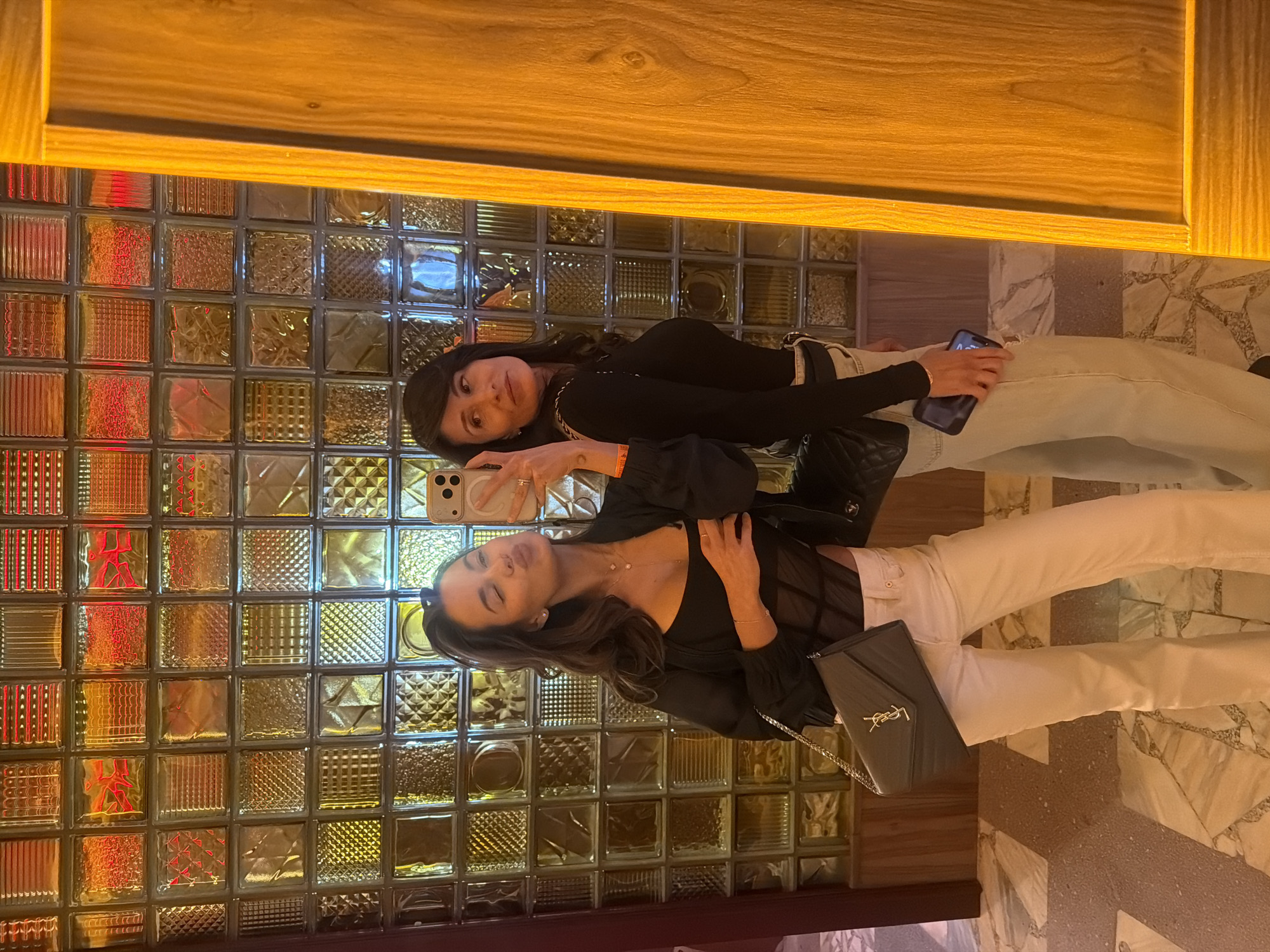 Two women are taking a selfie in front of a glass block wall. The woman in the foreground is wearing a black top and white pants. She is holding a phone in her right hand and is looking at the camera. She has a purse hanging on her left side. The woman in the background is wearing a black shirt and light blue pants. She is holding a phone in her right hand. The location may be a restaurant or bar with decorative glass blocks. There is no evidence that this location is in Krasnoyarsk, Russia.