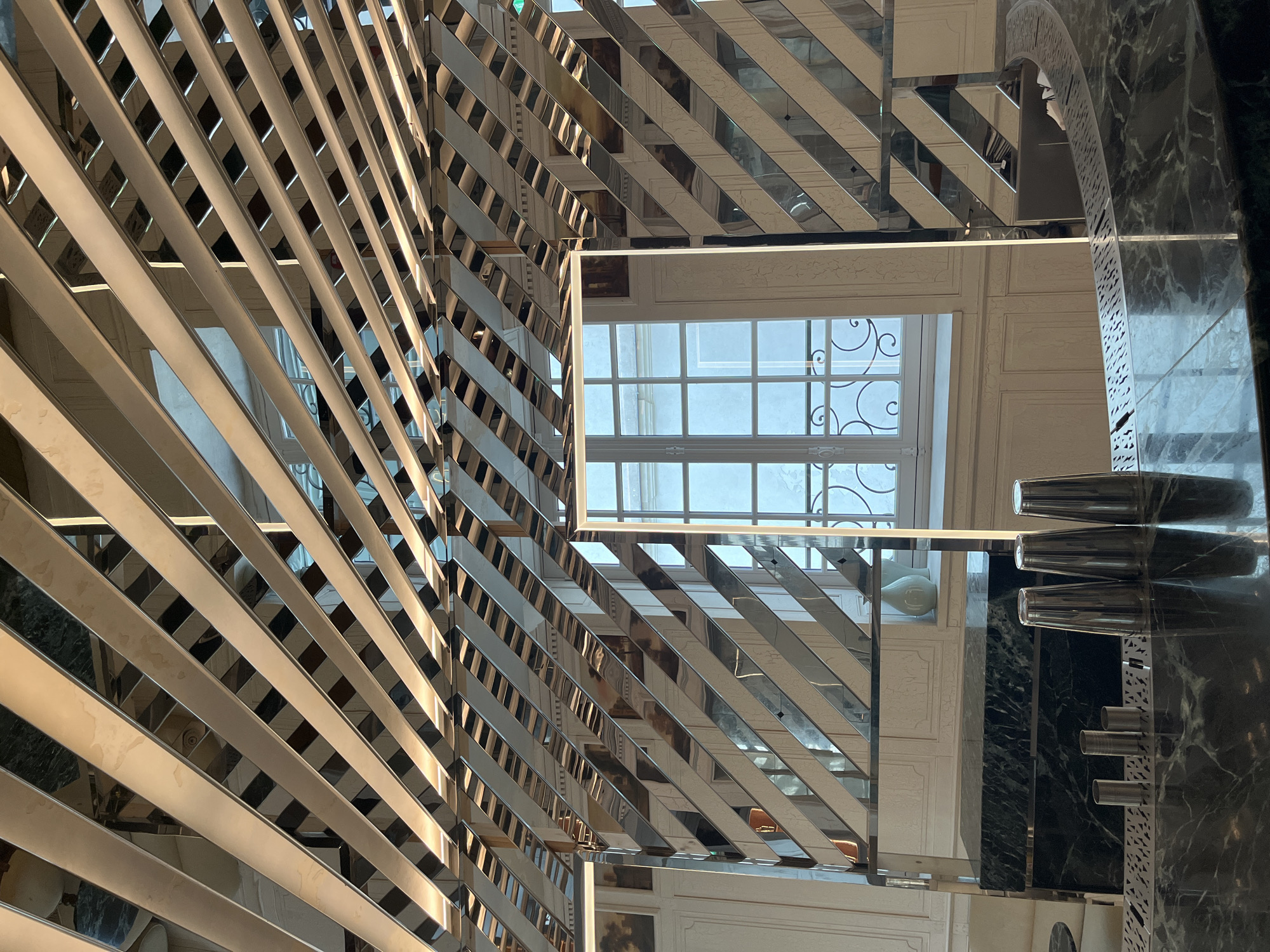 The image depicts a mirrored and slatted architectural detail in a Parisian setting. Light is diffused through a window, creating a pattern of light and shadow on the interior. The window, visible through the latticework, features decorative ironwork. On the right side, a dark marble surface reflects cocktail shakers and other bar equipment. The overall effect is one of refined elegance and reflects Parisian architecture and design.