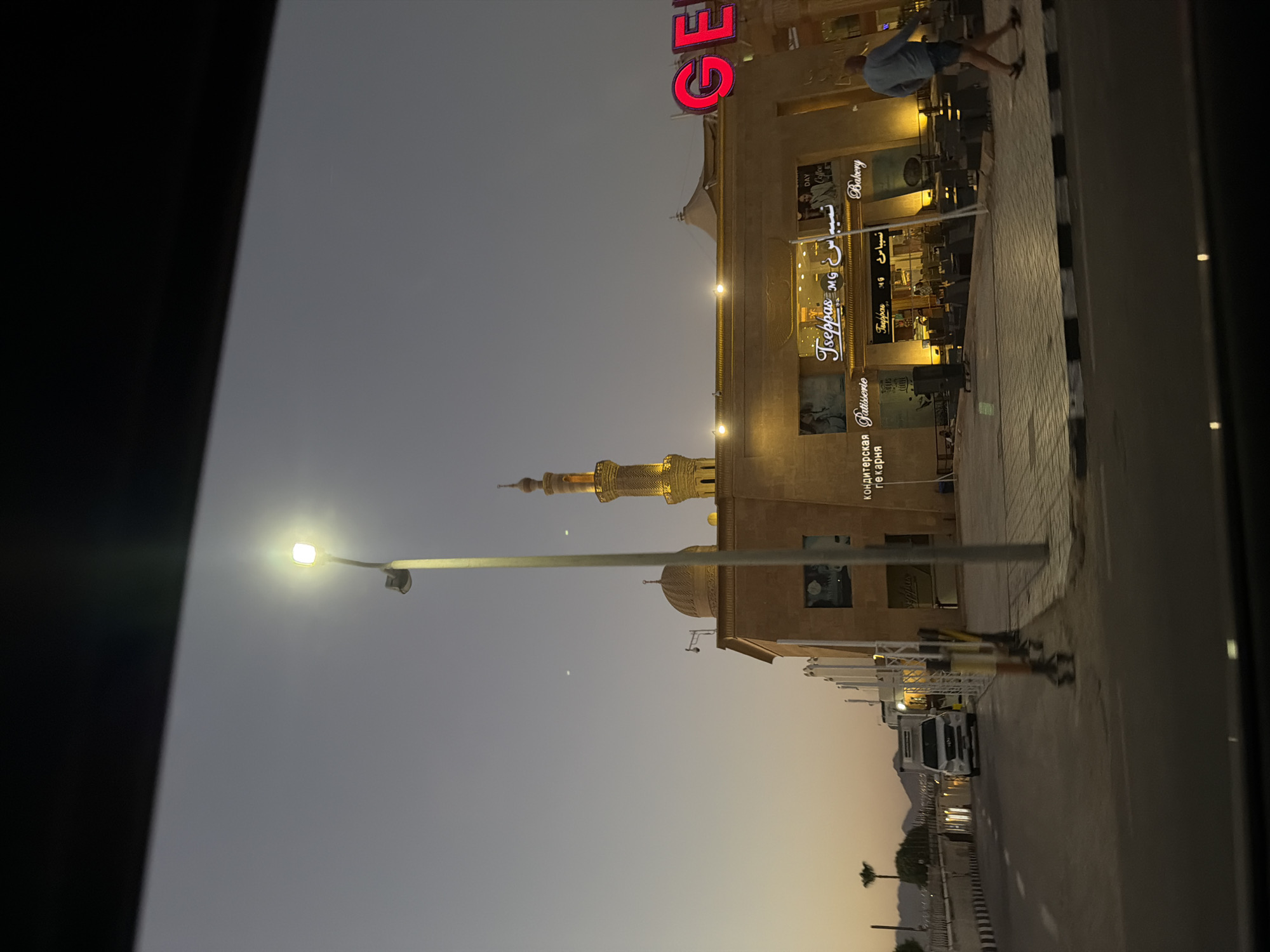 This is a photograph taken at dusk in Sharm El Sheikh, Egypt, likely on a street with commercial establishments. The sky is a soft gradient of twilight colors, from deep blue at the top to a lighter orange-pink near the horizon. A prominent street lamp with a bright, illuminated bulb is visible in the left half of the frame.
In the background, a building with a light-colored facade is illuminated by warm interior lights. The building features architectural elements reminiscent of local styles, including a dome and a tall, slender minaret topped with a golden spire. The minaret is also lit up, highlighting its intricate design.
Signage on the building indicates a business named "Tseppas MG," with text in both English and Arabic that appears to refer to a patisserie and bakery. The words "Patisserie" and "Bakery" are clearly visible, along with the Arabic equivalent. Outside the establishment, tables and chairs are arranged, suggesting an outdoor seating area.
On the right side of the building, large red neon letters spell out "GE," possibly part of a larger sign.
A man is walking away from the building towards the right side of the frame. He is wearing a light blue shirt and dark shorts. Further to the left, a white van is parked on the side of the road, and in the distance, mountains can be seen silhouetted against the fading light. The road in the foreground has a black and white striped curb. The overall impression is that of a bustling commercial street in the evening.