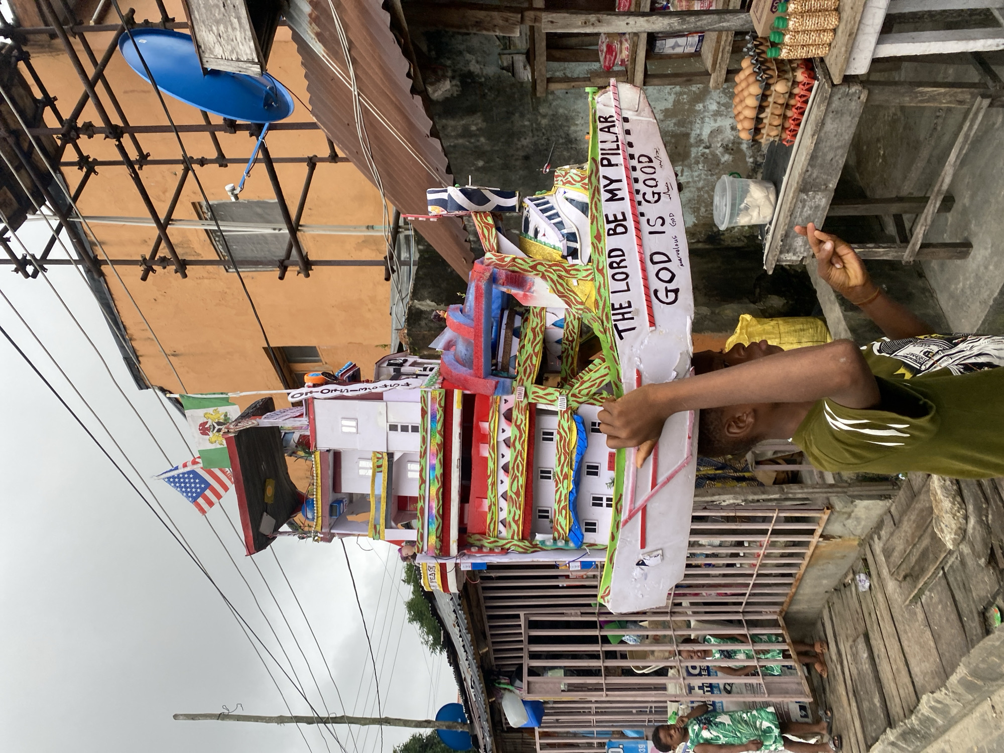 In an outdoor urban environment, a young Black male wearing a green t-shirt with white stripes carries a large, multi-story model of a boat on his head. The boat is elaborately decorated with colorful painted details and features flags, including the American flag and a Nigerian flag. Text on the boat reads, "THE LORD BE MY PILLAR" and "GOD IS GOOD". He is walking on a wooden boardwalk next to a metal fence. Behind the fence, two young Black females in green floral dresses are visible. The background includes buildings with orange and corrugated metal exteriors, scaffolding, a blue satellite dish, and a street-level market stall displaying trays of eggs and bottled goods. Power lines crisscross the overcast sky.