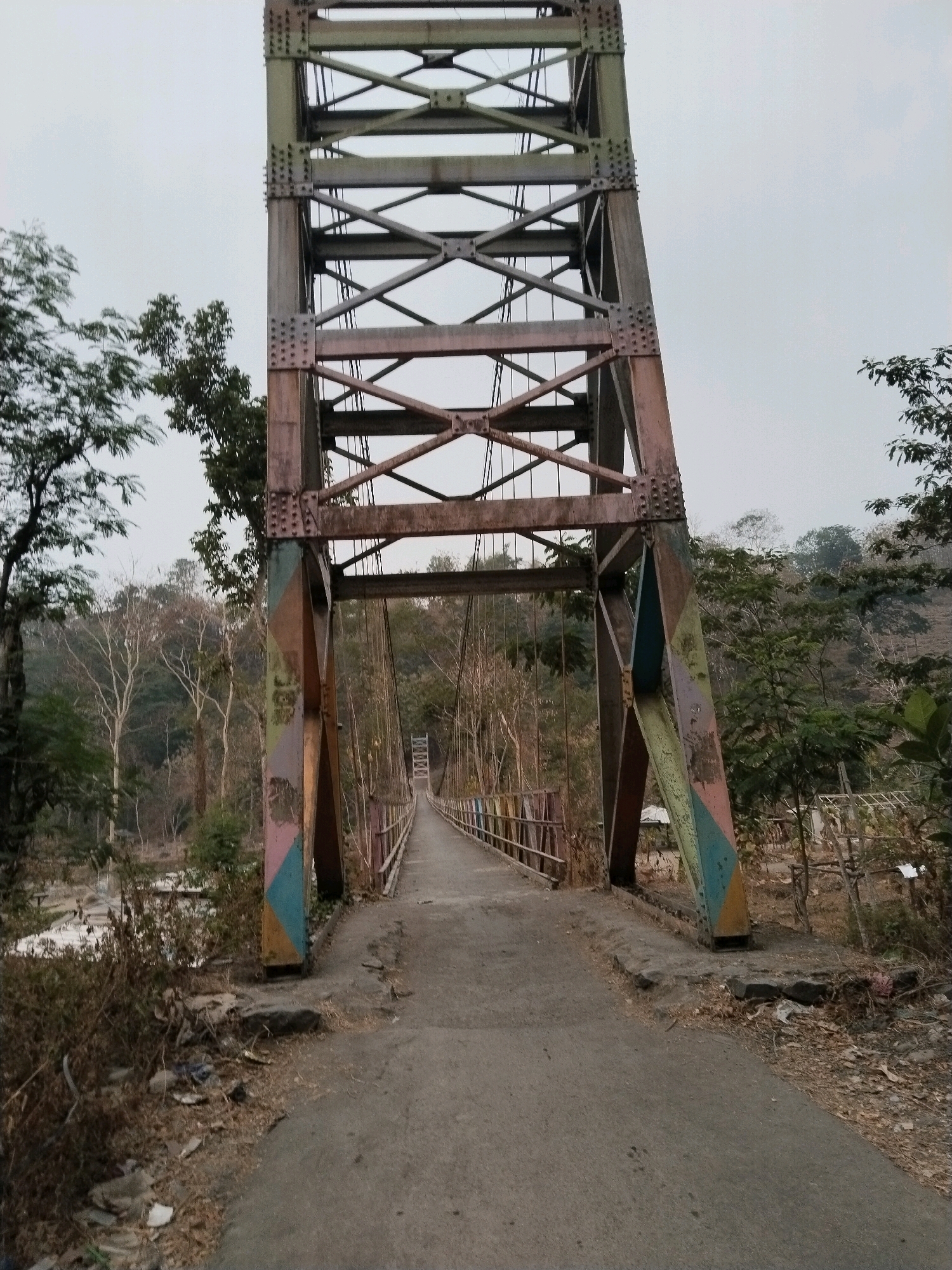 The image shows a suspension bridge with a tall, steel truss structure at its entrance. The bridge is made of metal and has a walkway that appears to be paved with asphalt. The supporting towers of the bridge are painted with colorful geometric patterns in shades of yellow, blue, pink, and green. The surrounding environment is lush and green, with trees and vegetation visible on either side of the bridge. The sky is overcast, suggesting a cloudy day. In the distance, the bridge extends across a body of water, disappearing into the trees and hills. The overall impression is one of a rustic and somewhat weathered but functional bridge in a natural setting. The location is identified as Sangkanjaya, Indonesia, suggesting it is a local bridge within that area.