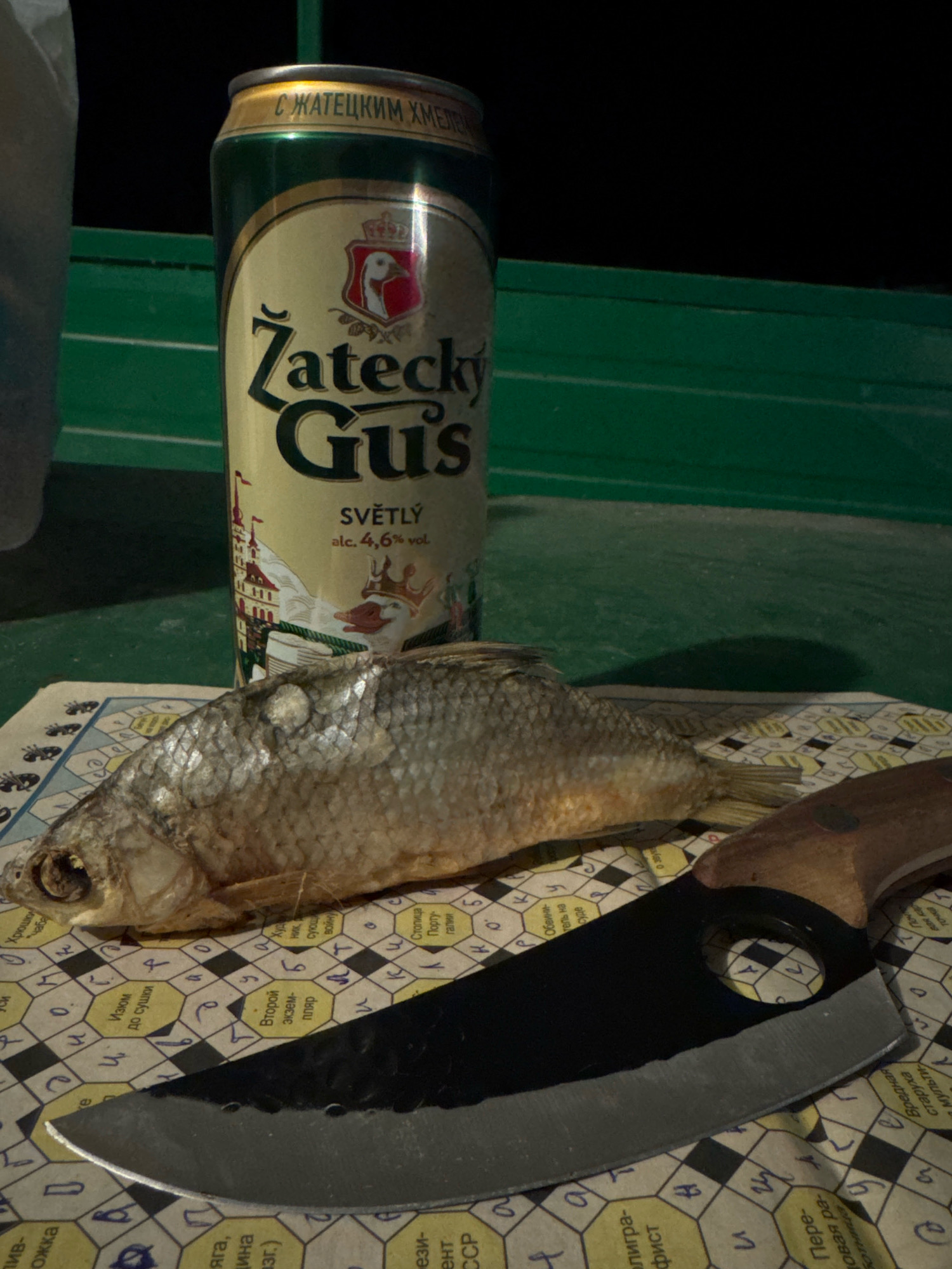 The image shows a still life of a dried fish, a can of Zatecky Gus beer, and a knife on a crossword puzzle. The fish is laid out horizontally, with its head facing left. The beer can is upright, with its label prominently displayed. The knife is positioned in front of the fish, with its blade facing right. The crossword puzzle serves as the background, with its grid of squares and letters visible. The scene is lit from above, casting shadows on the table. There is no indication of the city of Koklaisay, Kazakhstan.