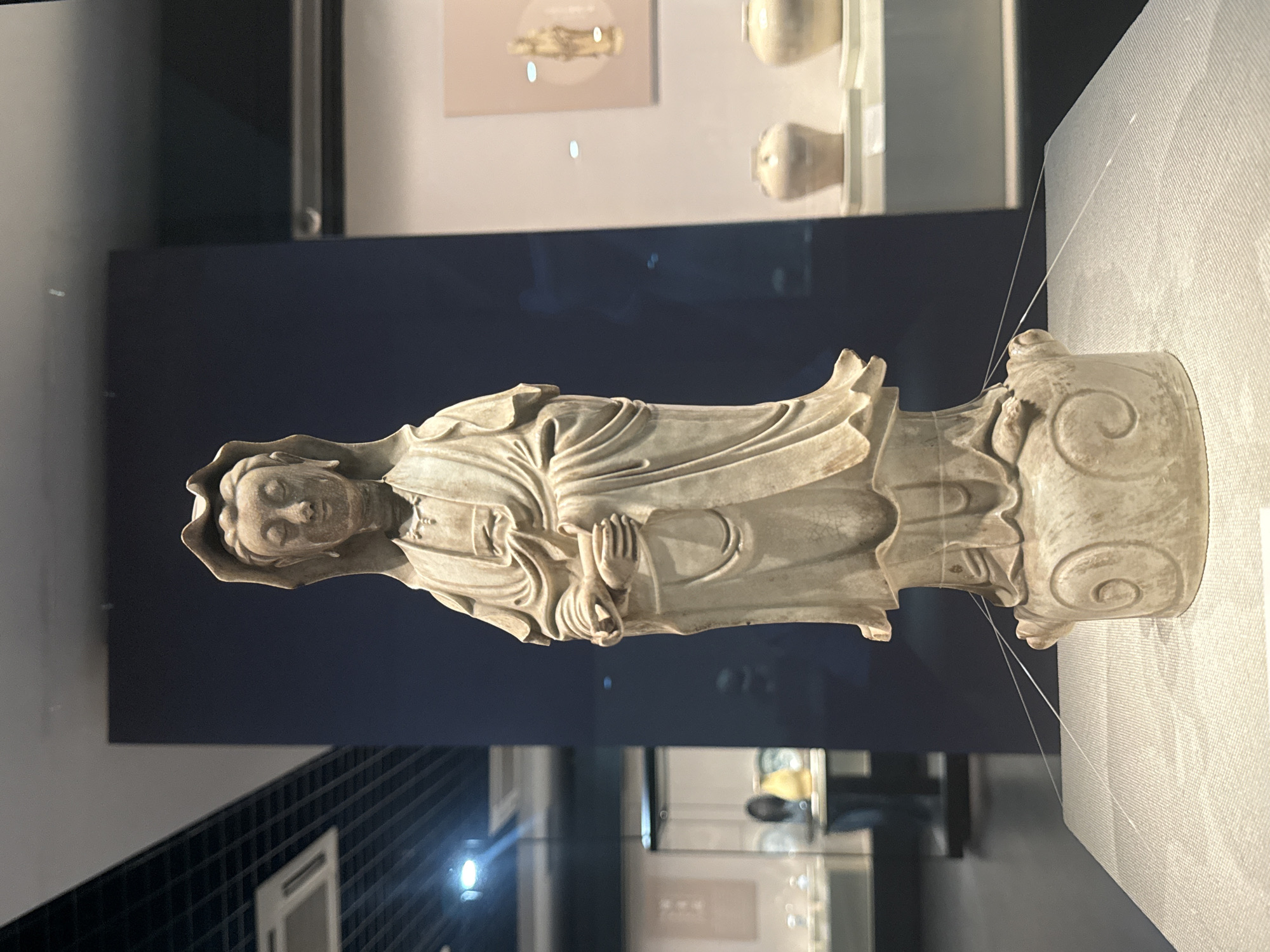 A weathered, light-colored stone statue, possibly of a religious figure, stands in a museum display. The statue is of a person in draped clothing with hands clasped near the chest. It is mounted on a base with scroll-like designs. The statue is under a suspended cable system. Behind the statue is a dark blue wall and reflective glass display cases. The floor has a black patterned design. The location is likely in a museum in Yuzhong District, China.