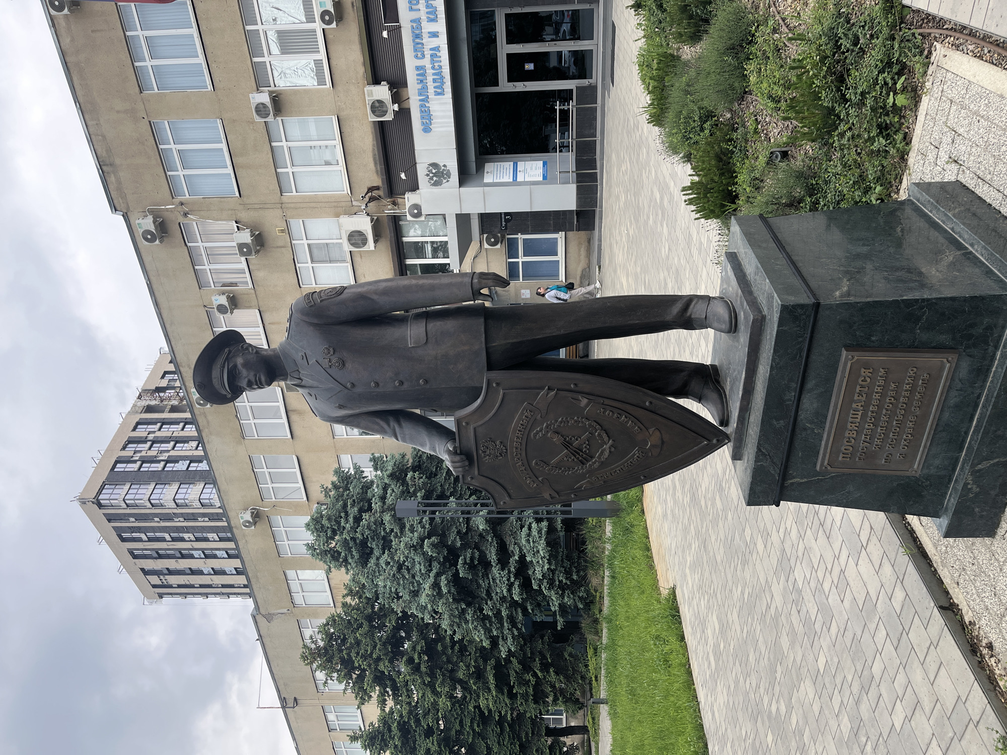 Here's a detailed description of the image:
**Scene Overview:**
The image shows a bronze statue of a uniformed officer holding a shield with a coat of arms. It's situated on a dark-colored stone plinth. Behind the statue is a modern building with many windows and air conditioning units on the exterior. A sign above the entrance of the building reads "ФЕДЕРАЛЬНАЯ СЛУЖБА ГОСУДАРСТВЕННОЙ КАДАСТРА И КАРТ" (FEDERAL SERVICE OF STATE CADASTRE AND CARTOGRAPHY). Trees are situated between the statue and the building.
**Specific Elements:**
* **Statue:** The statue is a man in a military-style uniform and cap, holding a shield with what appears to be a coat of arms on it.
* **Plaque:** A bronze plaque on the plinth reads "ПОСВЯЩАЕТСЯ государственным инспекторам по использованию и охране земель" (Dedicated to state inspectors for the use and protection of land).
* **Building:** The building is a multi-story structure with a symmetrical window arrangement and multiple air conditioning units attached to the facade. The sign identifies it as the Federal Service of State Cadastre and Cartography.
* **Vegetation:** There are evergreen trees between the statue and the building, along with some grassy areas.
* **Location:** The statue is located in the city of Krasnodar, Russia.
* **Weather:** The sky appears overcast.
**In summary, the image depicts a statue dedicated to land inspectors outside a building housing the Federal Service of State Cadastre and Cartography in Krasnodar, Russia.**